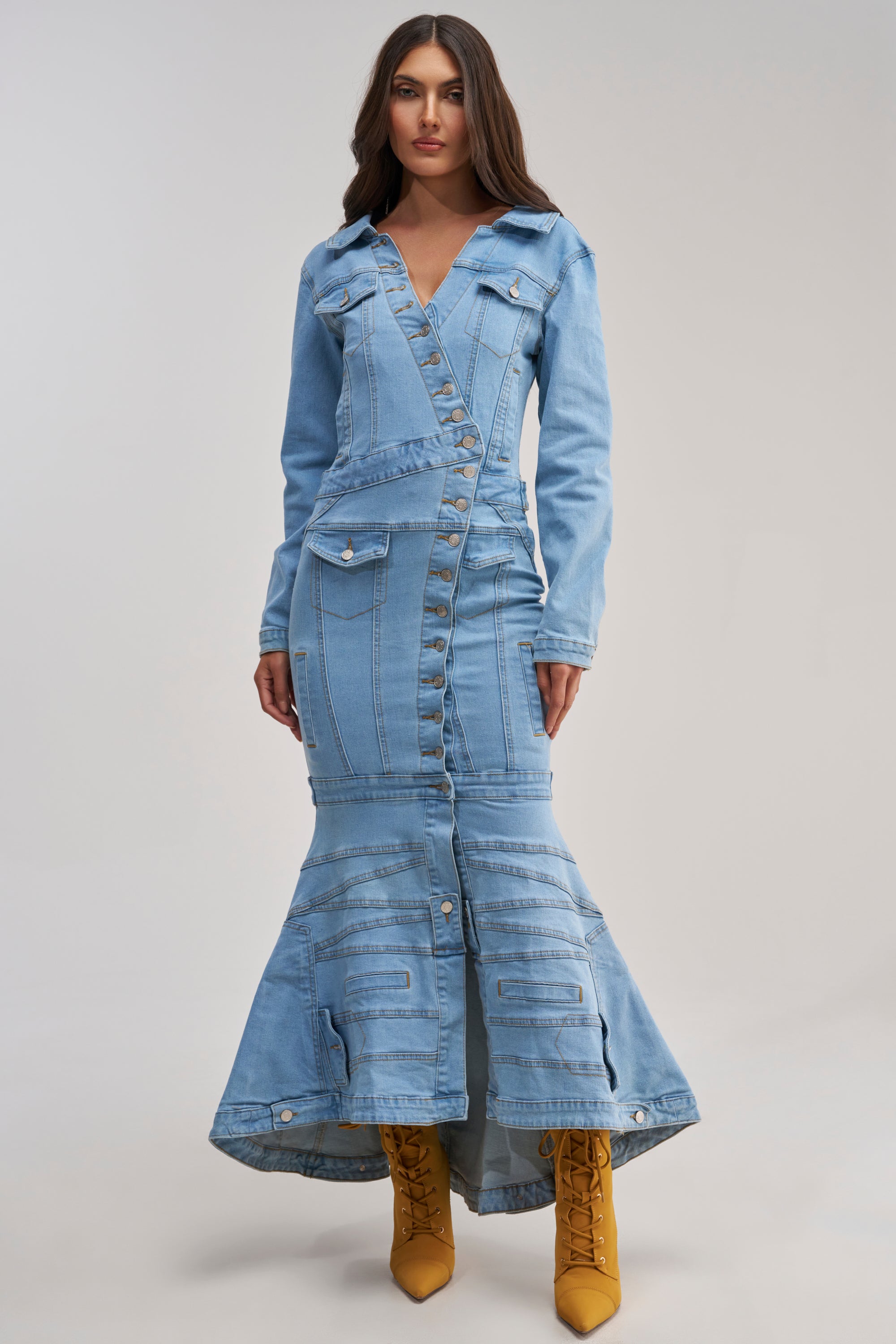 A woman wears the TRAPEZE BUTTON DOWN DENIM MAXI DRESS in a light wash with long sleeves, button details, and a mermaid skirt. She stands facing forward, hair loose, yellow lace-up boots standing out against a plain background.