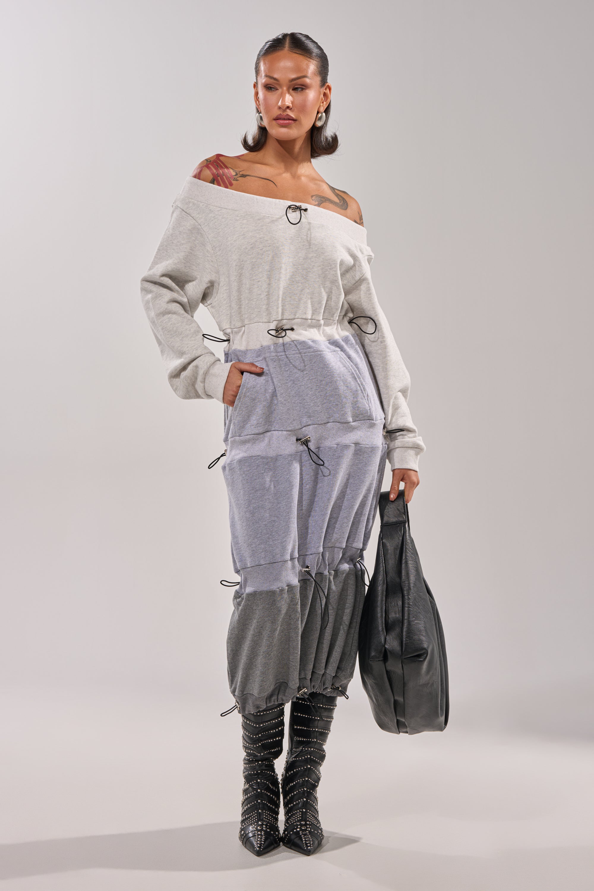A woman with long dark hair wears the TIED UP RIGHT NOW MAXI DRESS, an off-the-shoulder sweatshirt maxi in patchwork grey with drawstring accents. She styles it with knee-high studded boots, a large black bag, hoop earrings, and shows her tattoos. 0