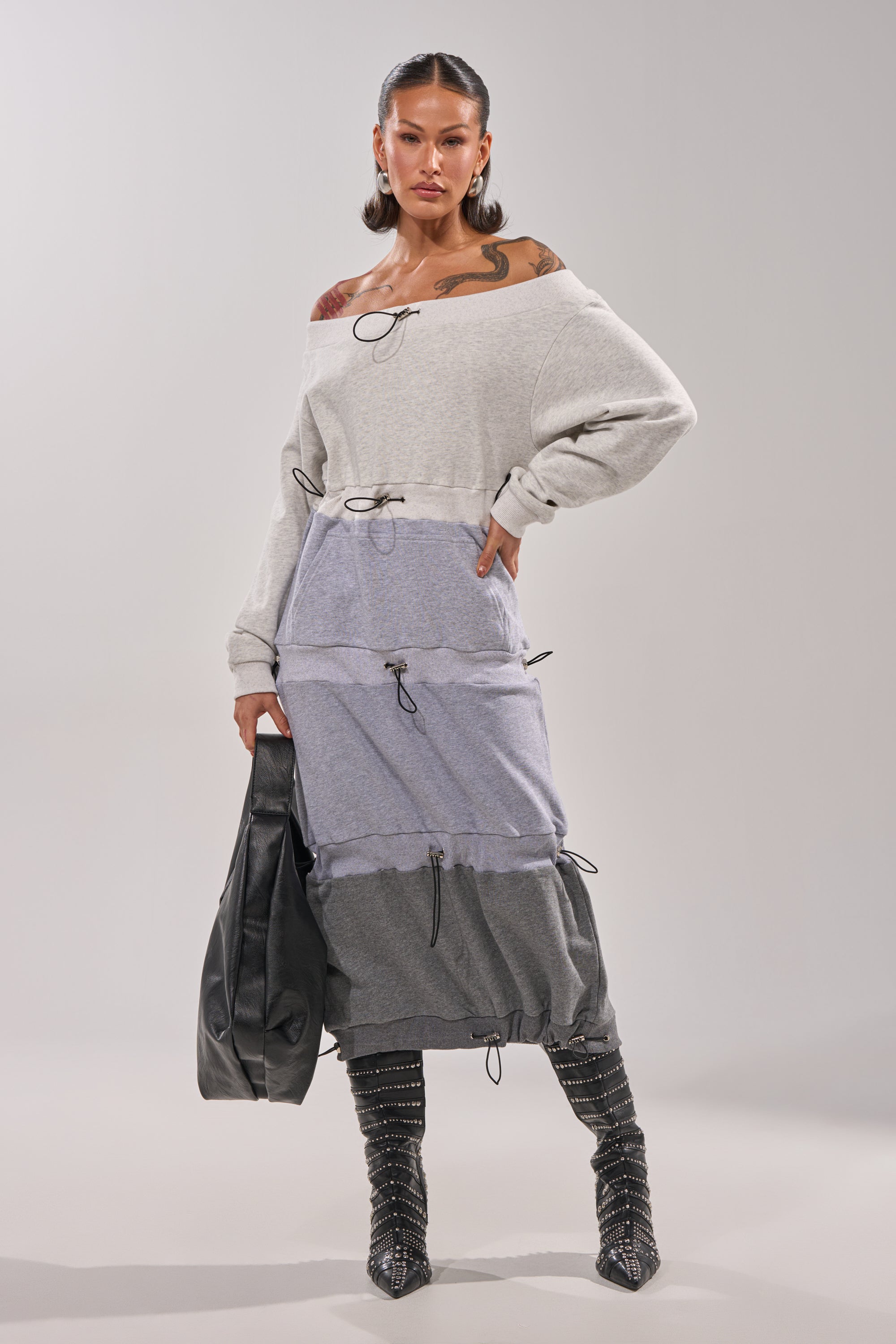 A woman models the TIED UP RIGHT NOW MAXI DRESS, an off-the-shoulder grey colorblock sweatshirt dress with drawstring details, paired with studded knee-high boots, bold earrings, and a black bag against a plain background. 1