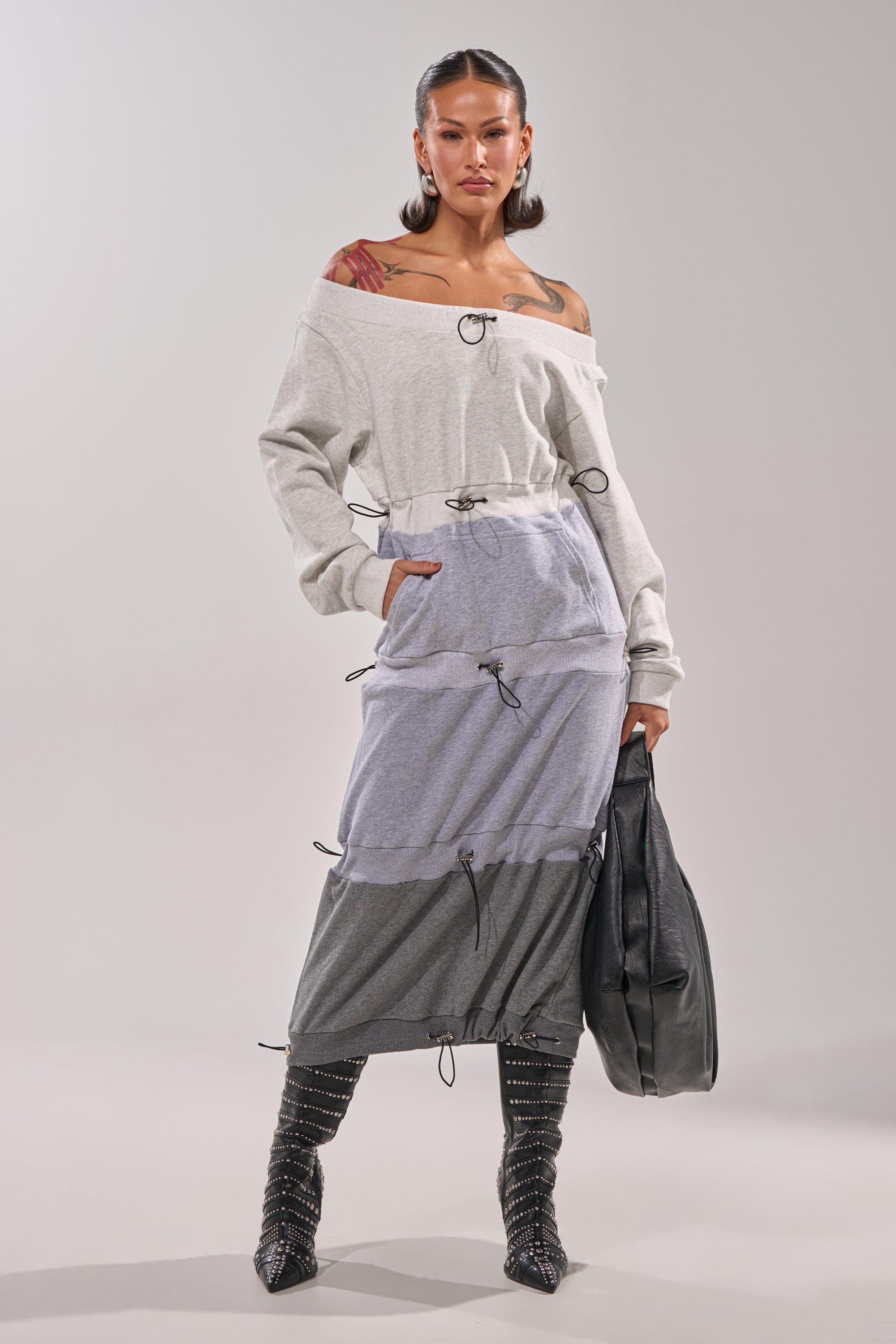 A woman wears the TIED UP RIGHT NOW MAXI DRESS—a colorblock, off-the-shoulder maxi with drawstring details—paired with black studded knee-high boots and a black bag, standing confidently against a plain background. 2