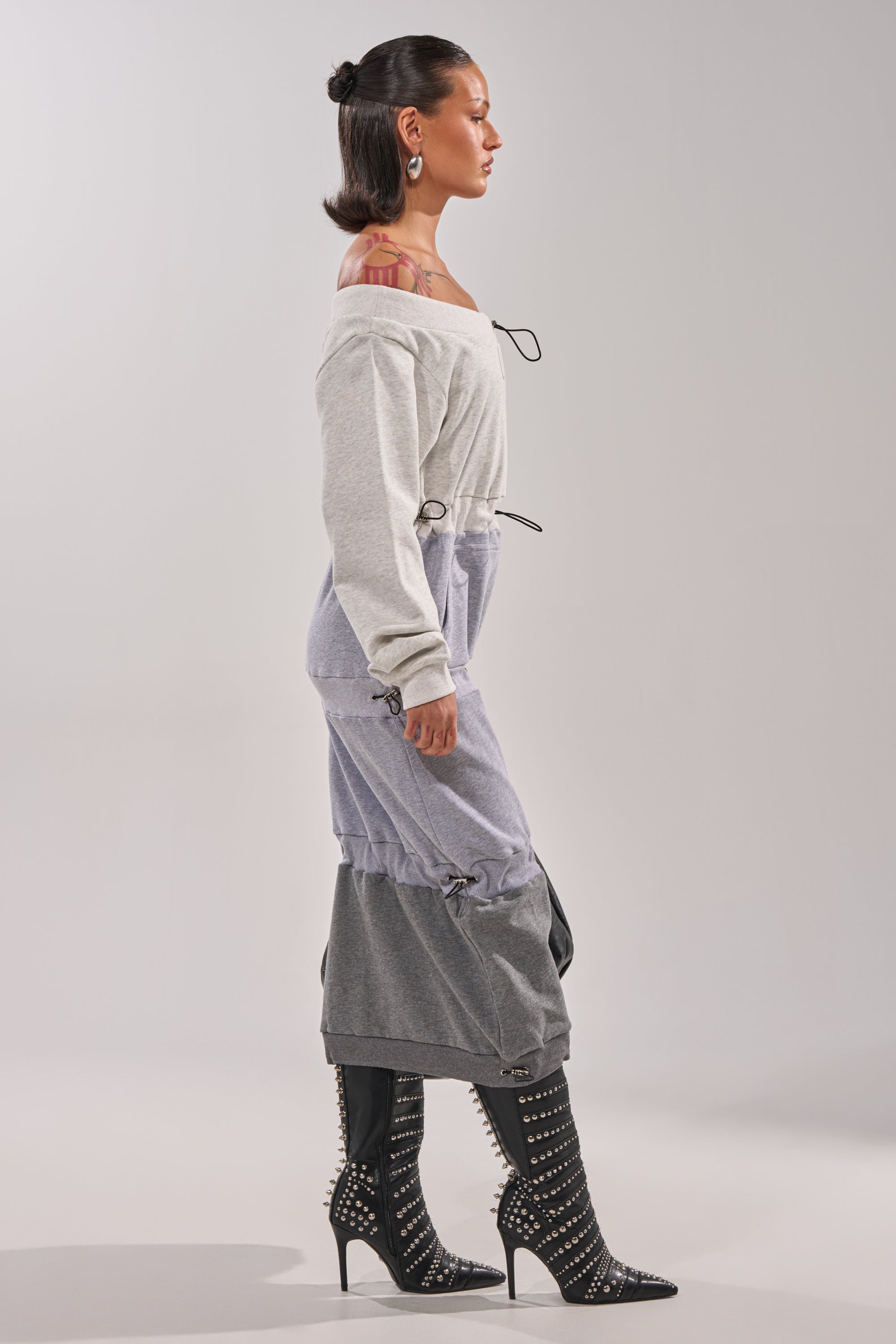A woman in profile wears the TIED UP RIGHT NOW MAXI DRESS—an off-the-shoulder, gathered sweatshirt maxi dress—styled with black studded high-heeled boots, slicked-back hair, and hoop earrings, set against a plain background. 3