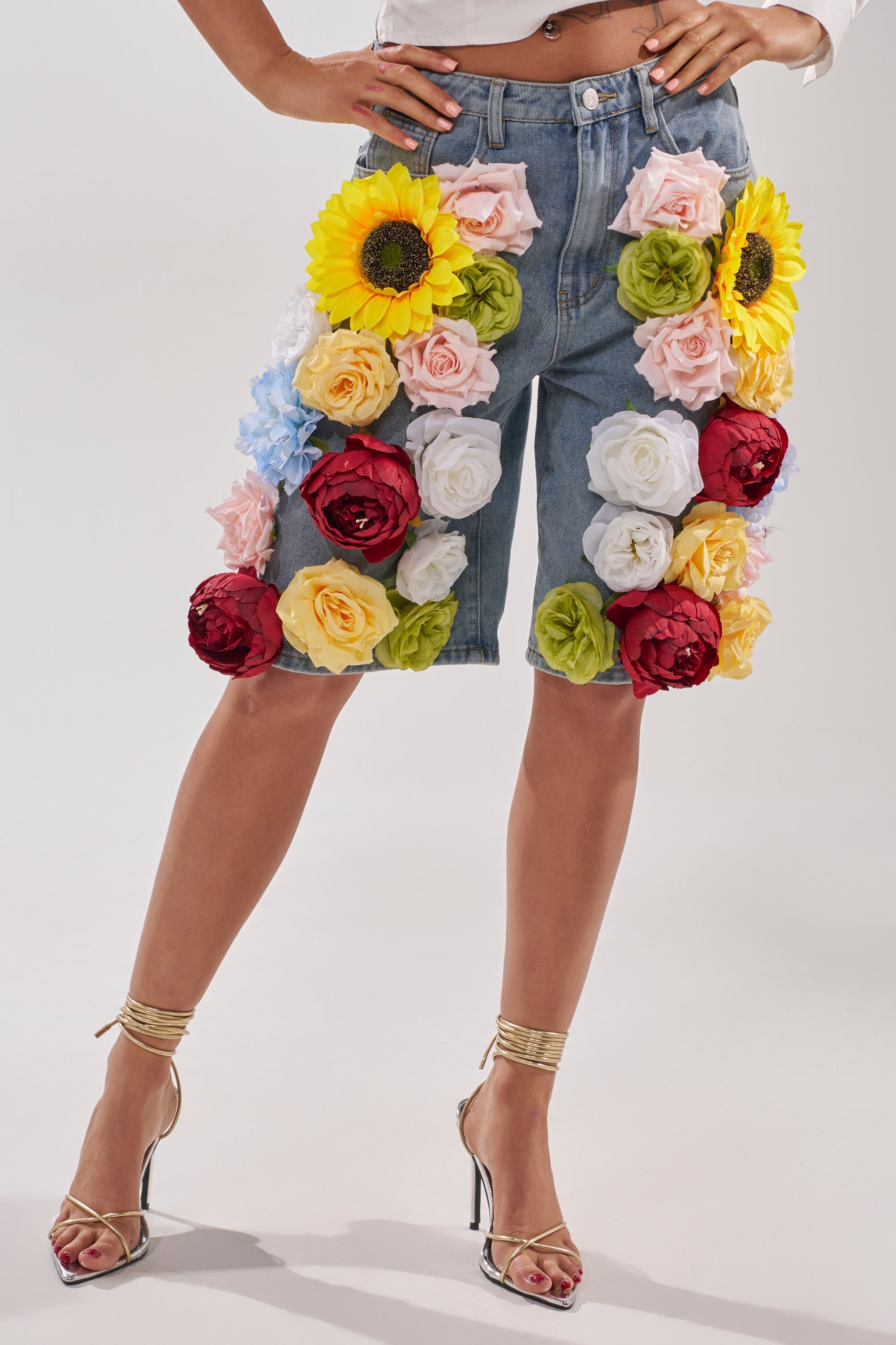 A person wearing FLOWER POWER BERMUDA SHORT, a high-rise denim short embellished with large artificial sunflowers, roses, and peonies in bold colors, paired with gold high-heeled sandals. 0