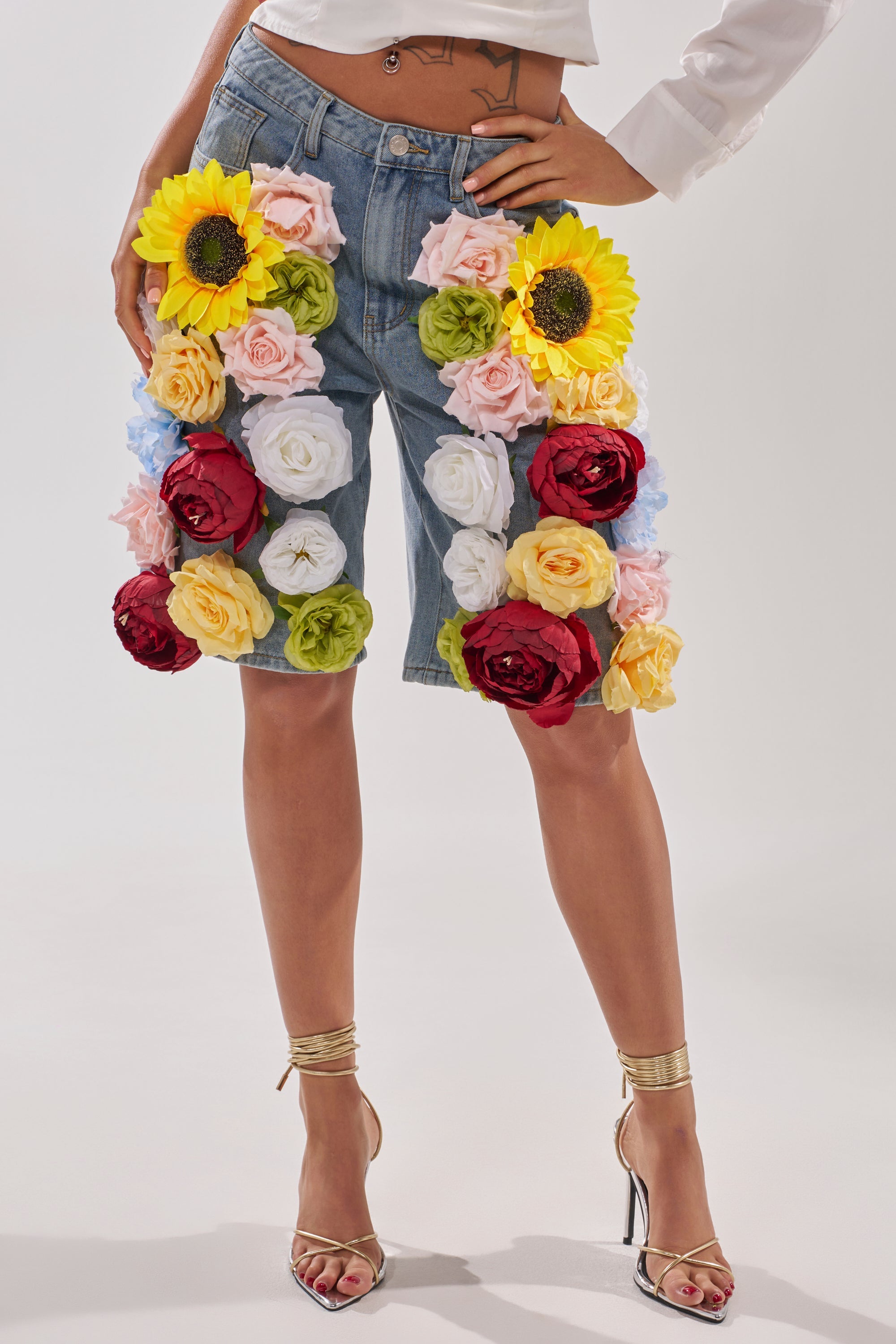 Wearing the FLOWER POWER BERMUDA SHORT, which features high-rise denim and bold sunflower and rose appliques, paired with gold heels and a crisp white shirt. 1