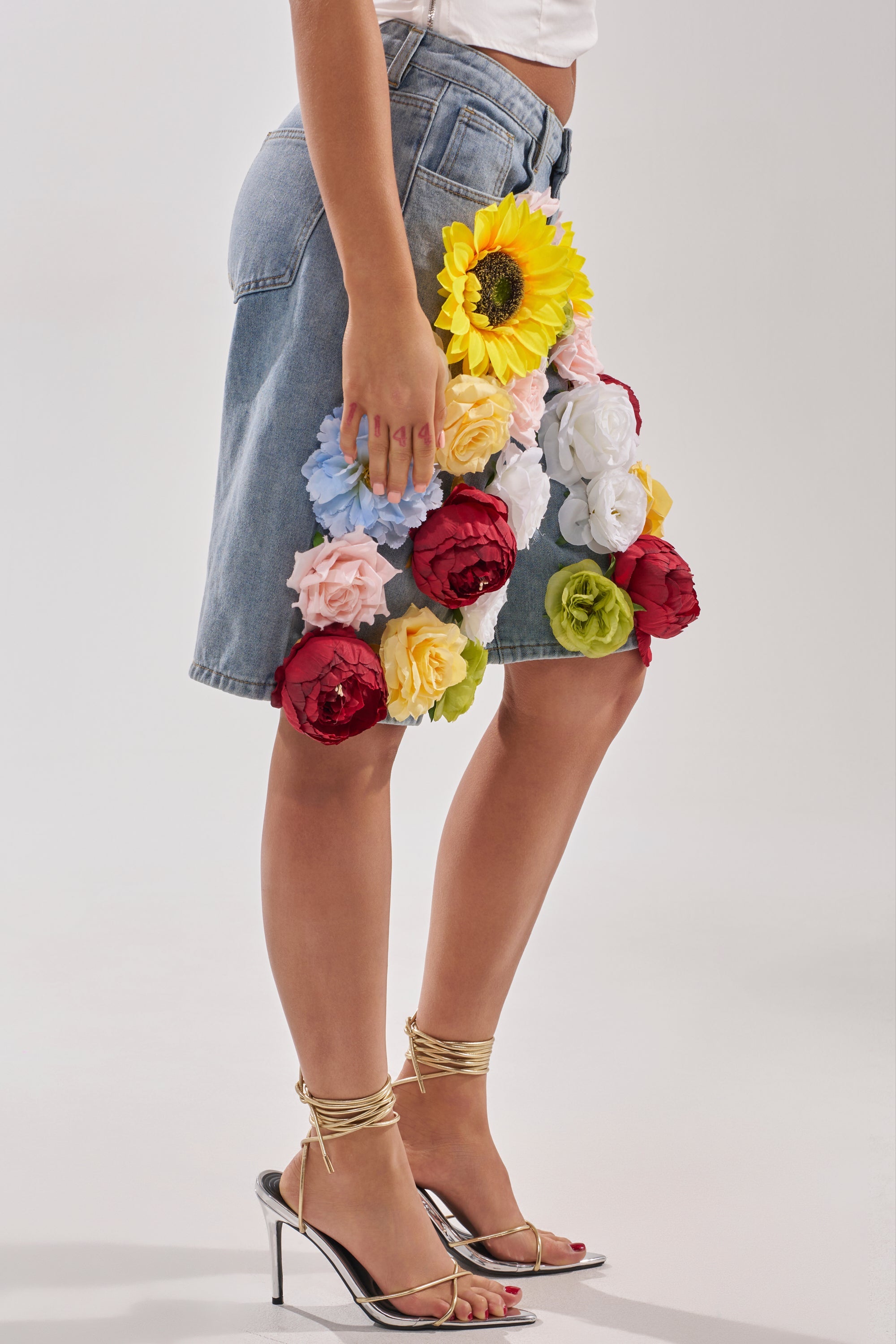 A person wears the FLOWER POWER BERMUDA SHORT, featuring bold, colorful floral appliques, paired with a white top and strappy high-heeled sandals against a plain light background. 2