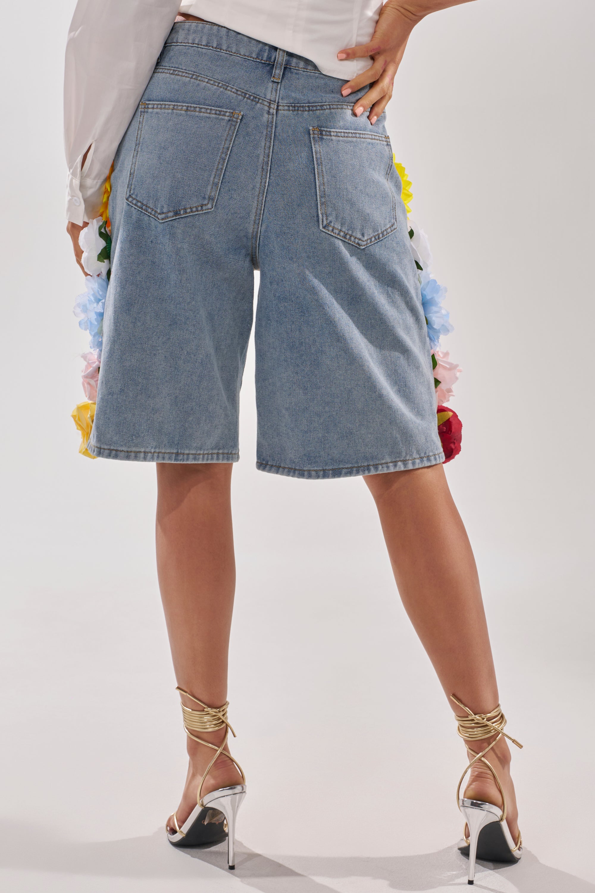 A person stands facing away, wearing FLOWER POWER BERMUDA SHORTS with floral appliques peeking from under the hem, paired with strappy gold heels and a white top. 3