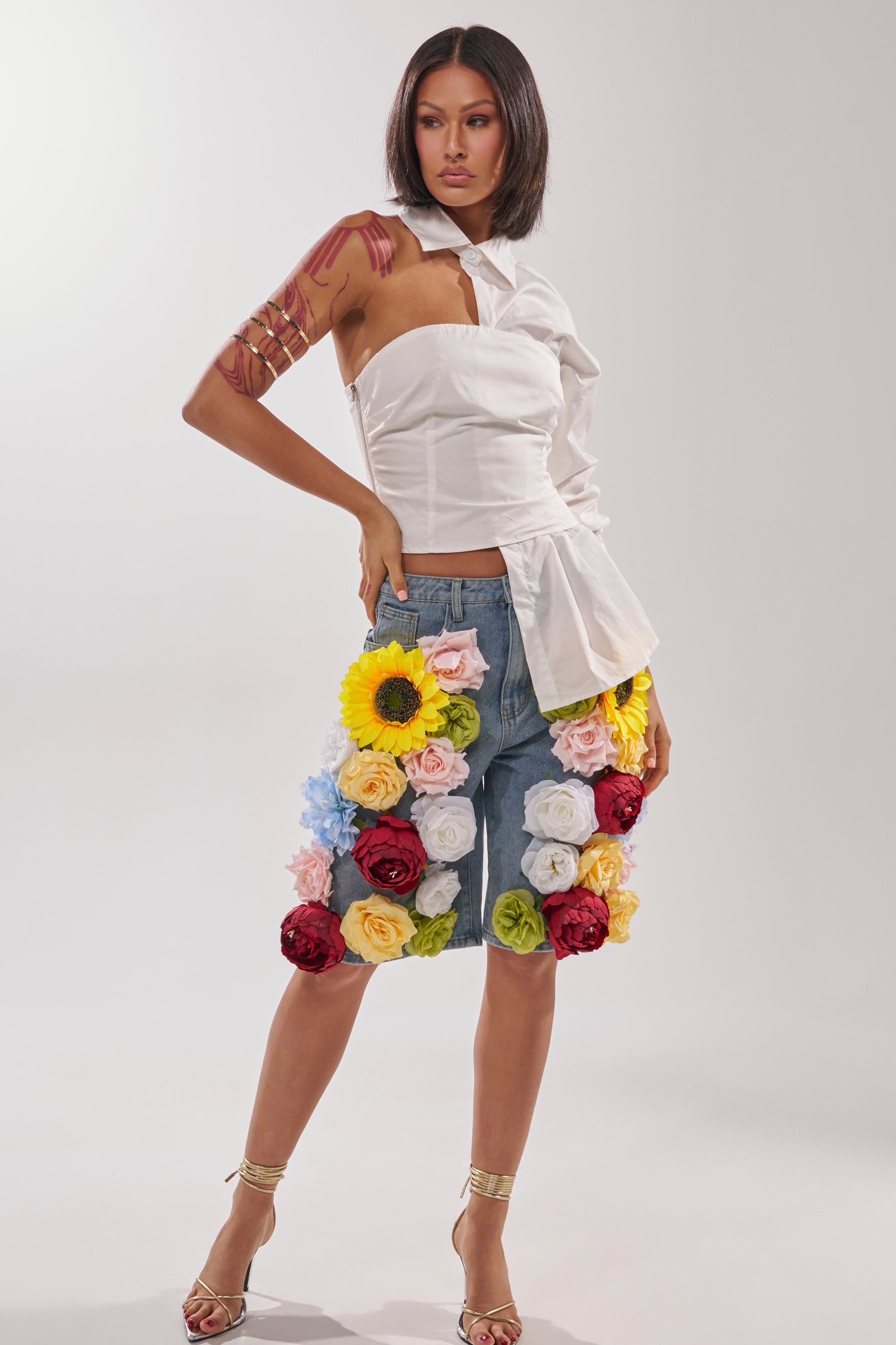 A woman poses in the FLOWER POWER BERMUDA SHORT, a high-rise denim style adorned with colorful floral appliques, paired with a white asymmetrical top and gold accessories against a plain light background. 4
