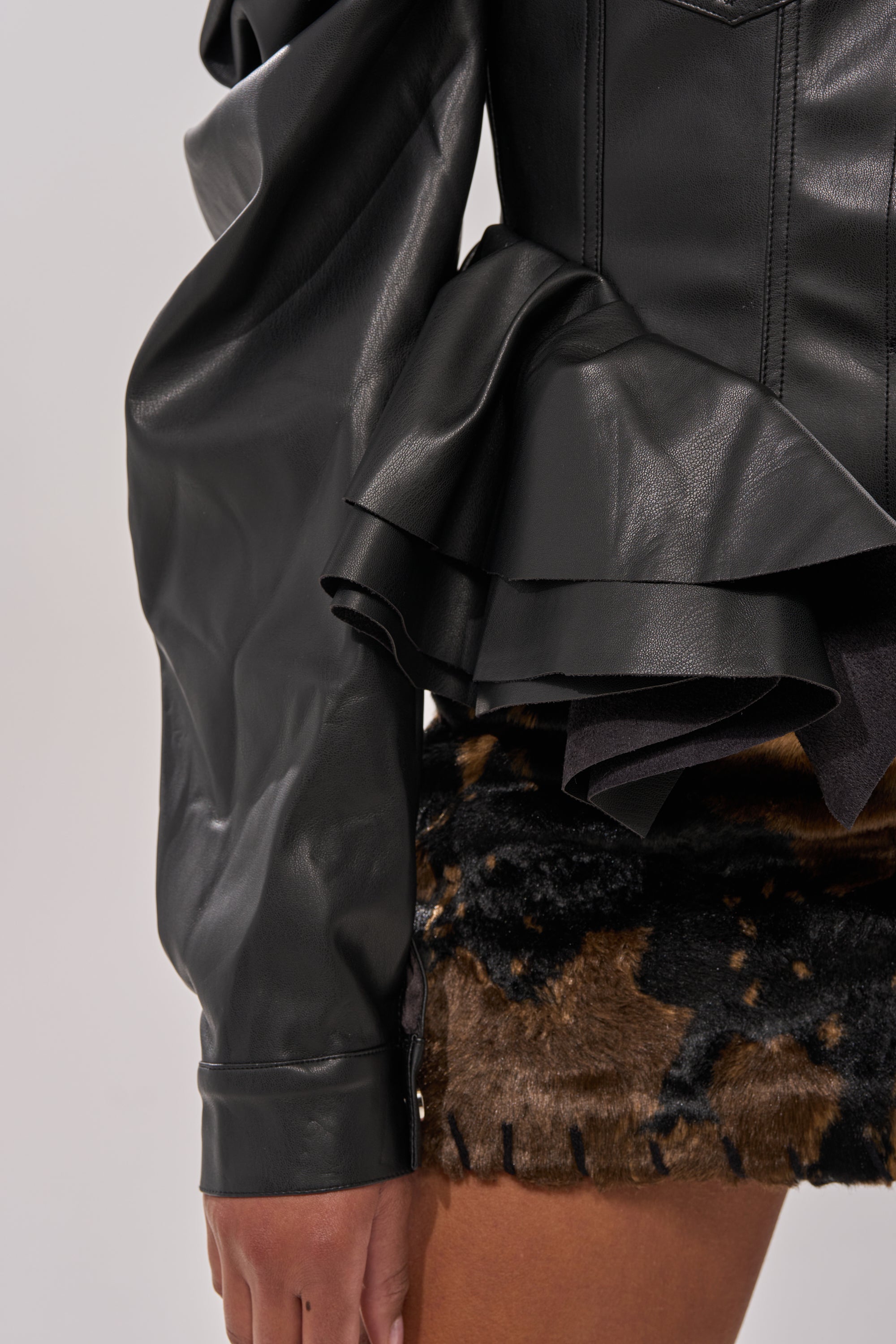 A close-up of a person wearing the NIGHT MOVES FAUX LEATHER JACKET, featuring puff shoulders and ruffled details, paired with a brown and black faux fur skirt. The focus is on the sleeve and waist area, with a hand partially visible. 9