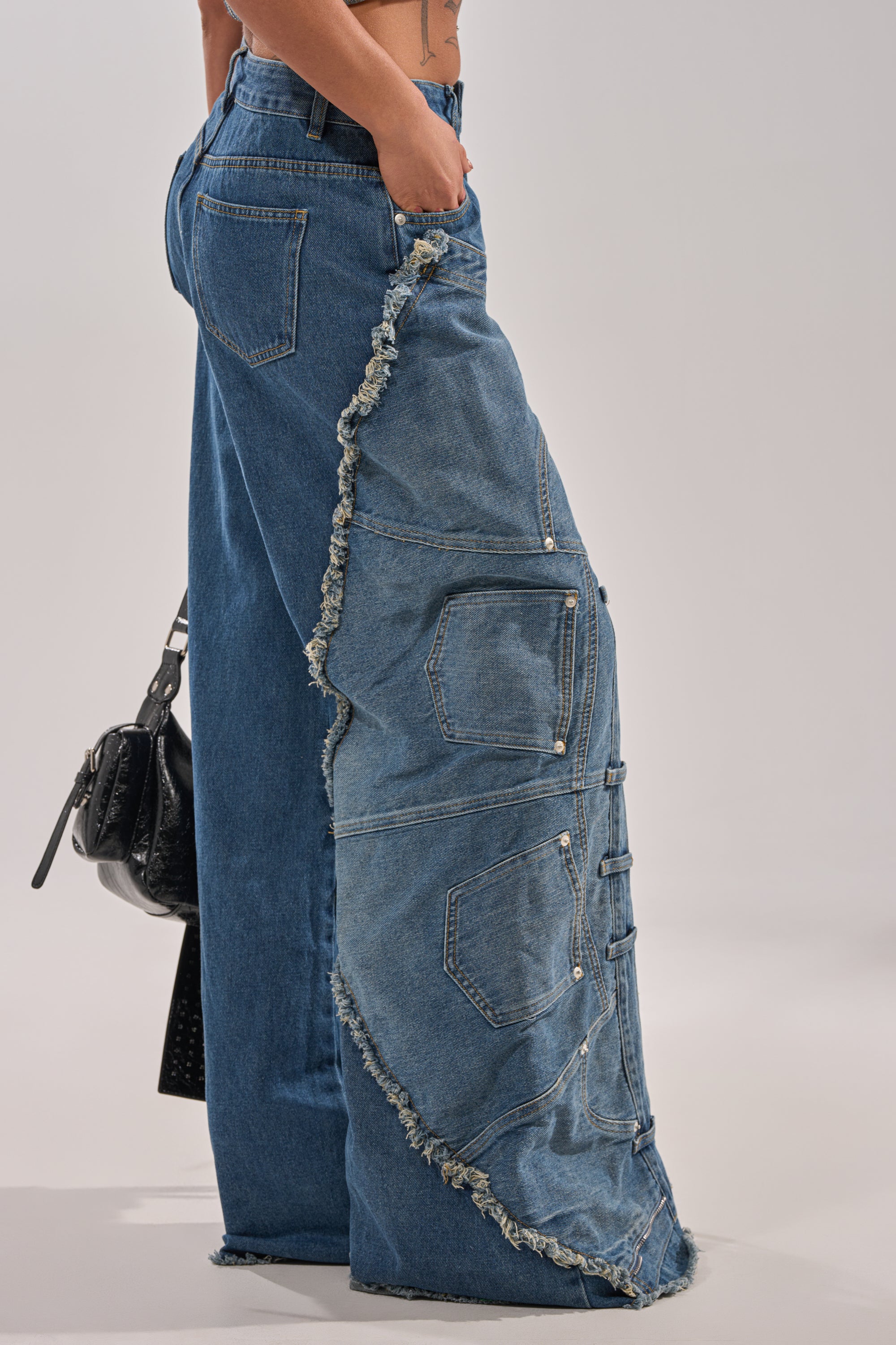 Wearing the MIXED MATCH RELAXED DENIM—streetwear-inspired wide-leg blue jeans with an asymmetrical frayed panel, pockets, and decorative stitching—paired with a black textured handbag. 3