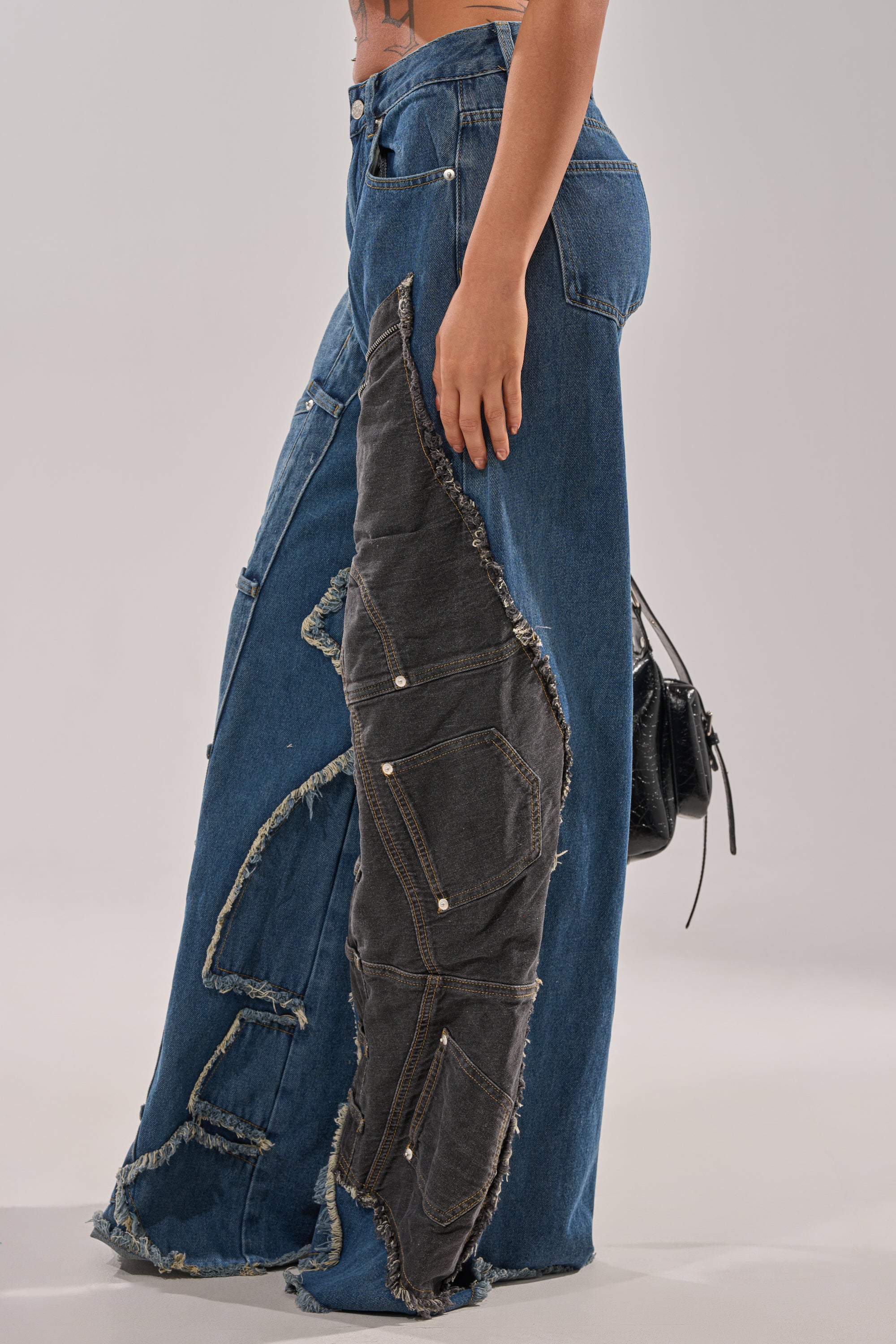 A person wears MIXED MATCH RELAXED DENIM featuring wide-leg blue jeans with large, frayed dark denim patches and multiple pocket outlines on the side, styled with a small black purse against a plain background. 4