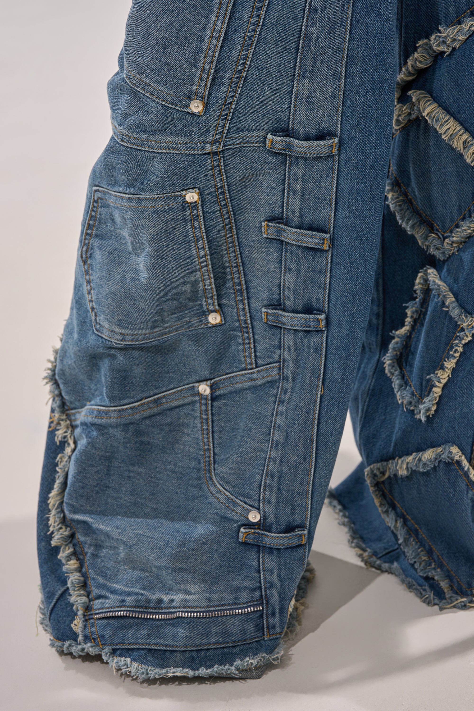 Close-up of MIXED MATCH RELAXED DENIM pants in blue patchwork with multiple pockets, exposed seams, frayed edges, extra belt loops, and decorative stitching for a textured streetwear vibe. 7