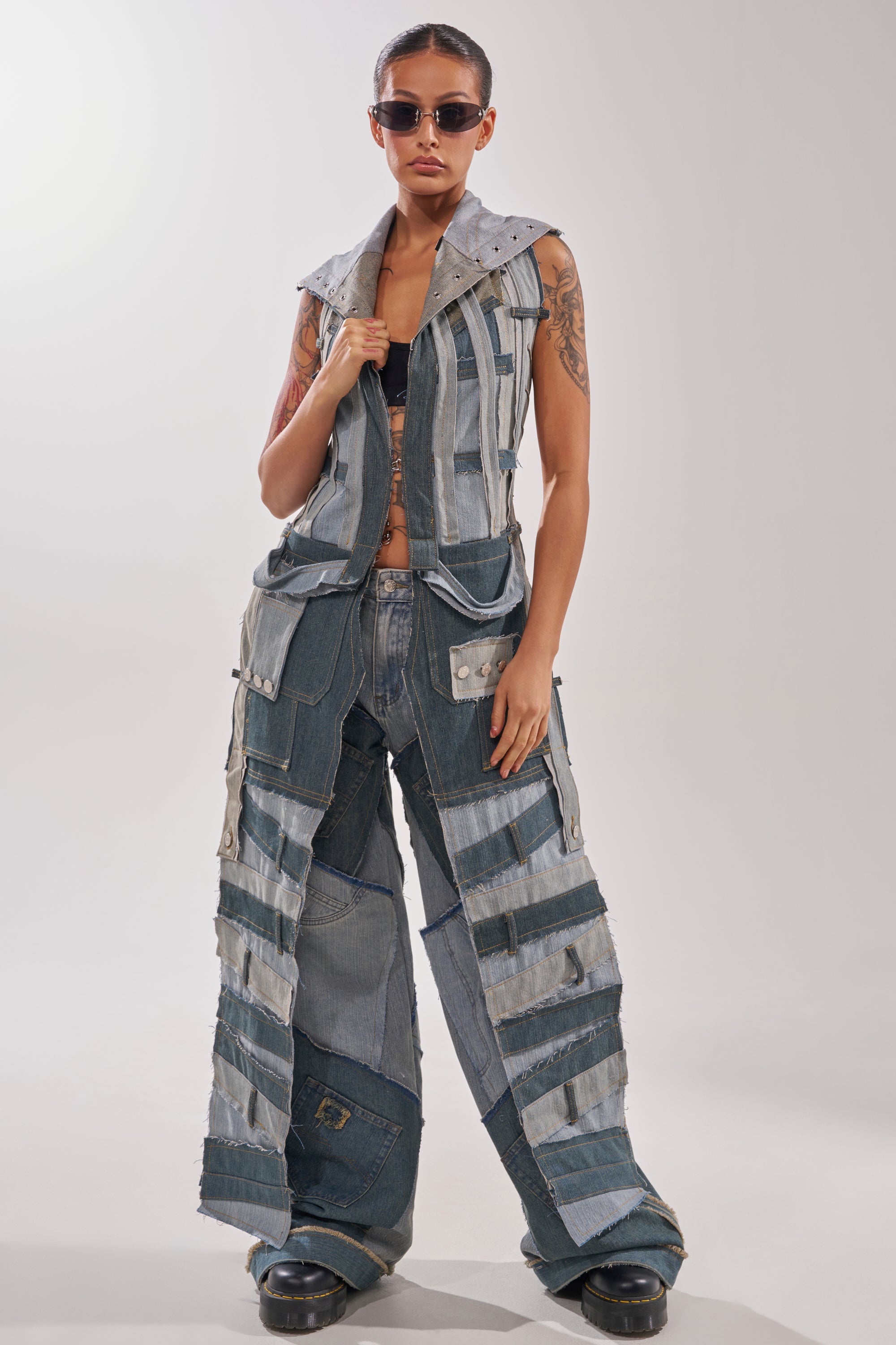 A woman stands confidently against a plain background in Y2K-style sunglasses, a sleeveless patchwork denim vest, ALL THAT WIDE LEG DENIM JEANS, chunky black shoes, and arm tattoos completing her bold look. 5