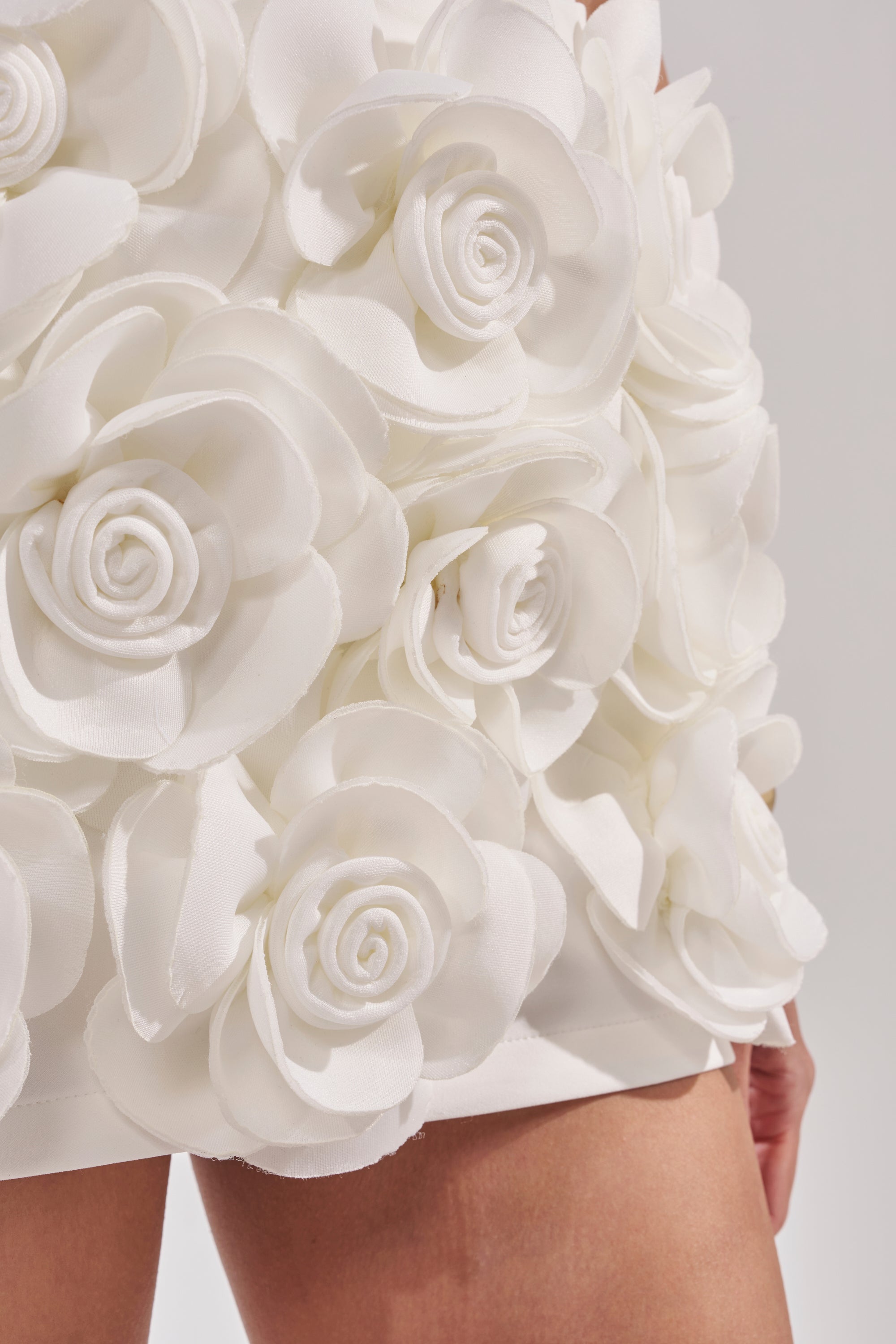 Close-up of the BRING ME FLOWERS MINI SKIRT, a white mini skirt featuring large 3D rosette appliqués, shown with a person's bare legs at the bottom. 8