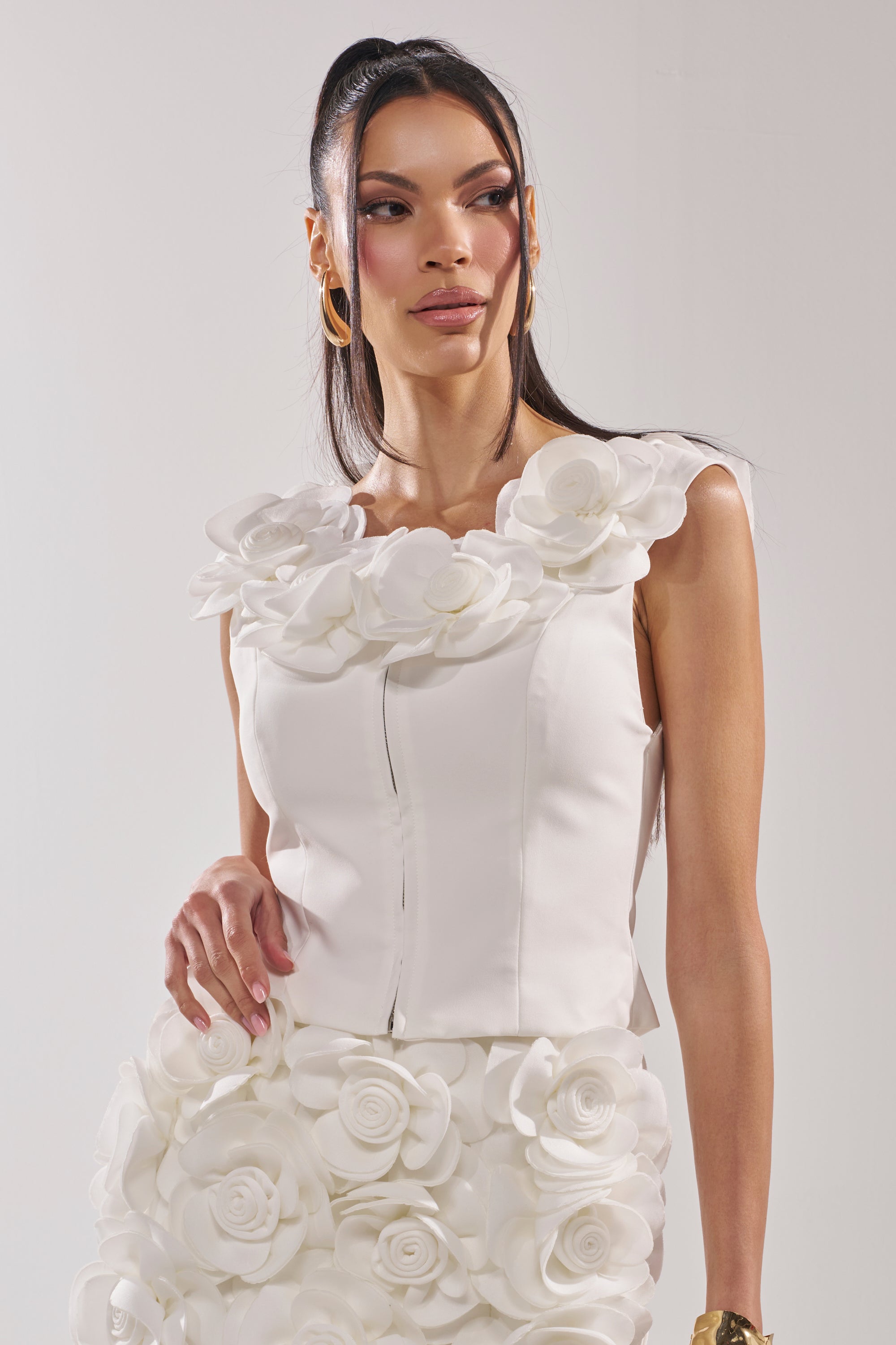 A woman with long dark hair wears the BRING ME FLOWERS BLOUSE, a sleeveless white top with a corset-style bodice and large 3D floral appliqués at the neckline. She pairs it with gold hoop earrings and looks to the side against a plain background. 0