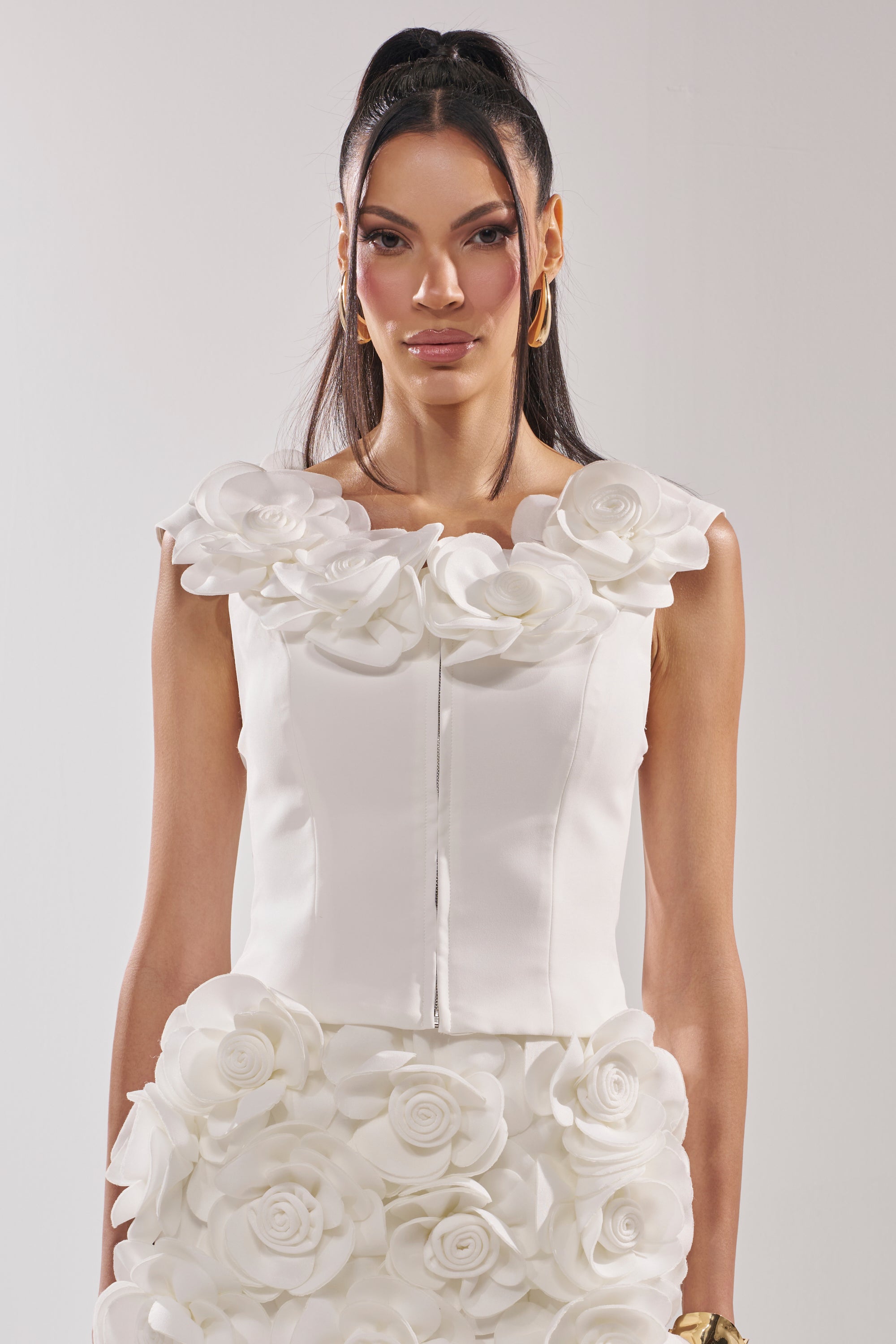A woman with long dark hair in a ponytail wears the BRING ME FLOWERS BLOUSE, a sleeveless white top featuring large 3D rosette appliqués around the neckline and hem. She completes her look with gold hoop earrings and a neutral expression. 1