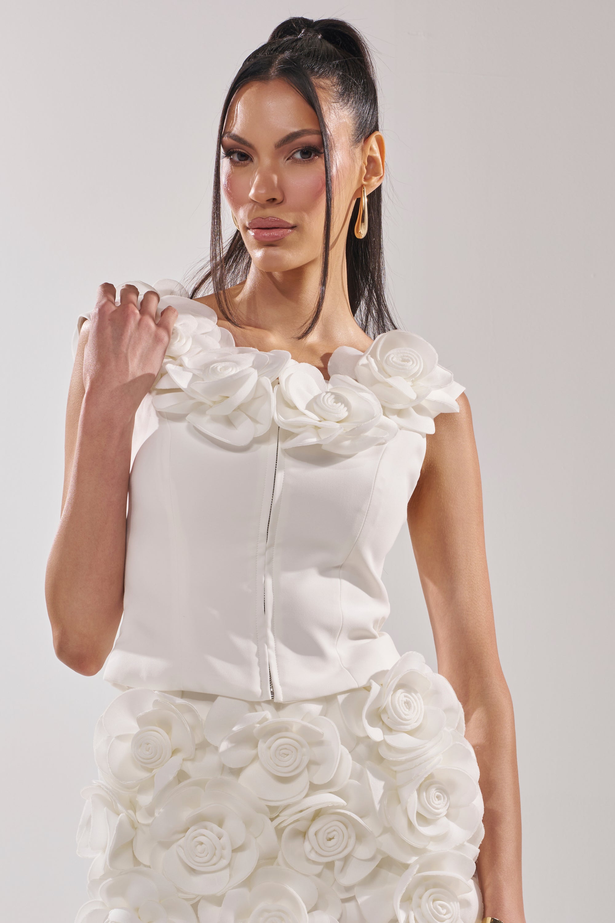 A woman with long dark hair in a high ponytail wears the BRING ME FLOWERS BLOUSE, a sleeveless white top with a corset-style bodice and bold 3D rosette appliqué, standing against a plain light background. 2