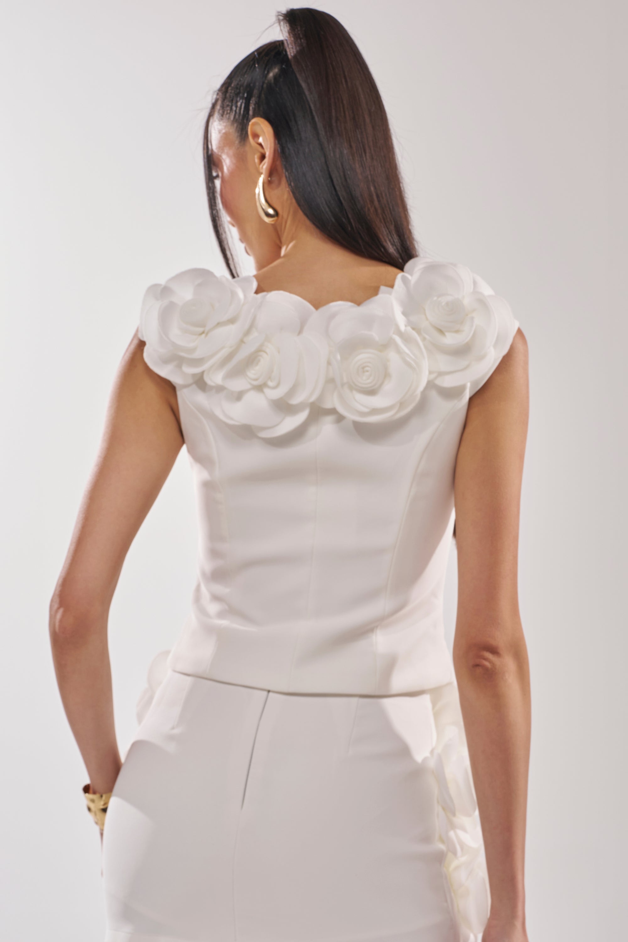 A woman with long dark hair and gold hoop earrings is seen from the back wearing the BRING ME FLOWERS BLOUSE, a white sleeveless top with 3D rosette appliqué at the neckline, paired with white high-waisted pants. 4