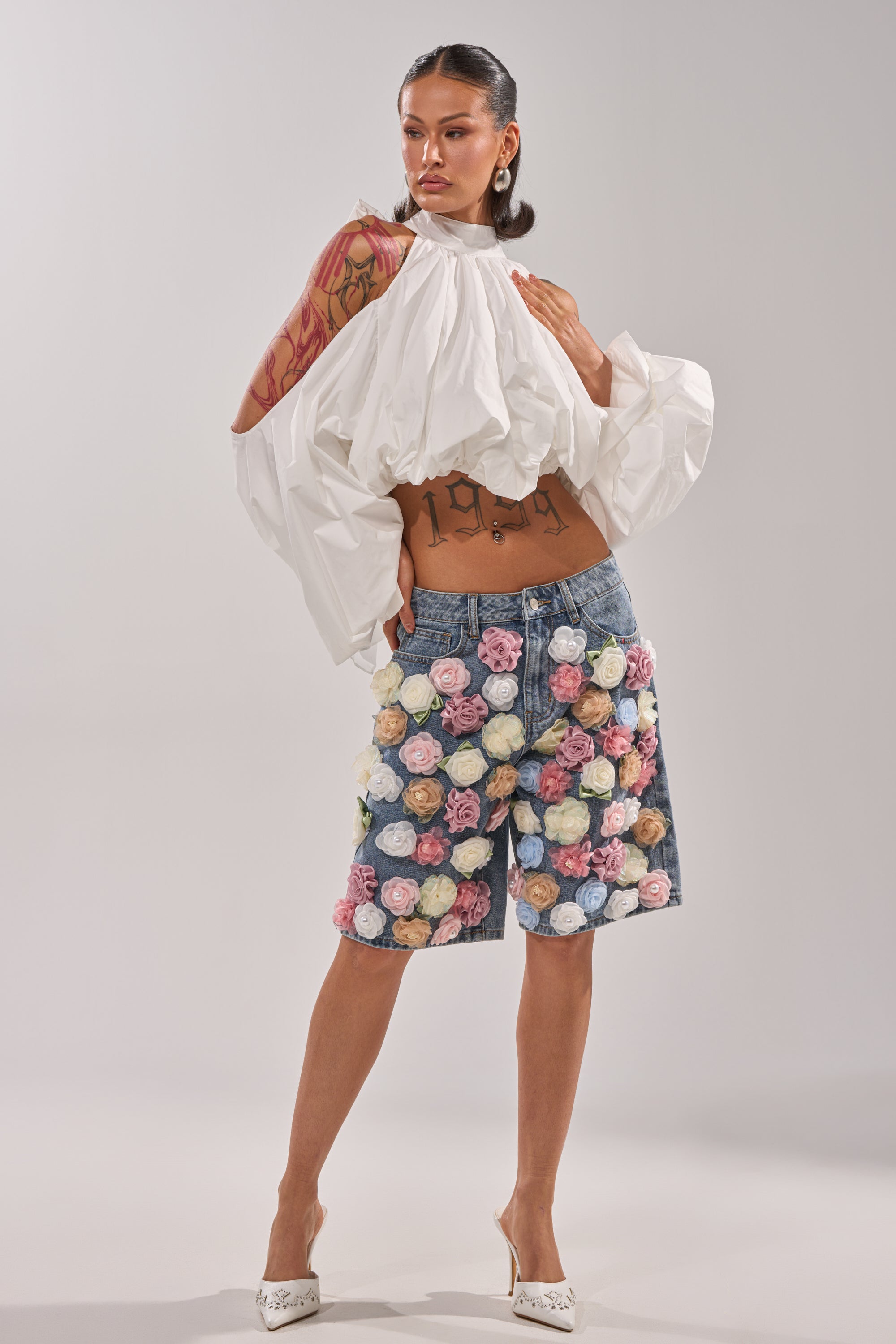 A woman models the IRIS FLORAL BERMUDA SHORT, high-rise denim shorts with 3D floral appliqués, paired with a white ruffled blouse and heels, revealing tattoos on her arms and torso. 0