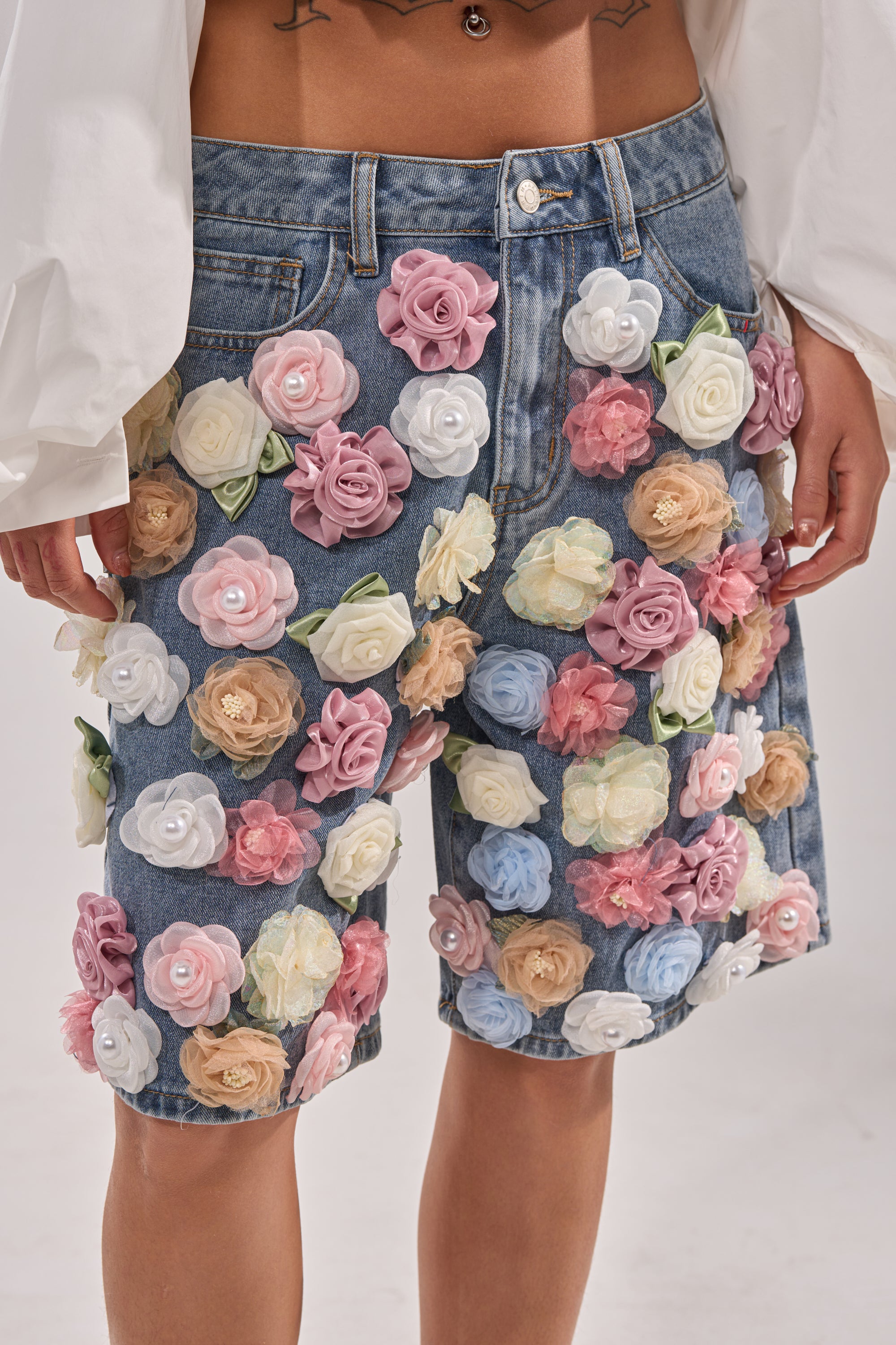 A person wearing the IRIS FLORAL BERMUDA SHORT, featuring high-rise denim and colorful 3D floral appliqués in pink, white, yellow, and peach tones, paired with a white long-sleeve top. 1
