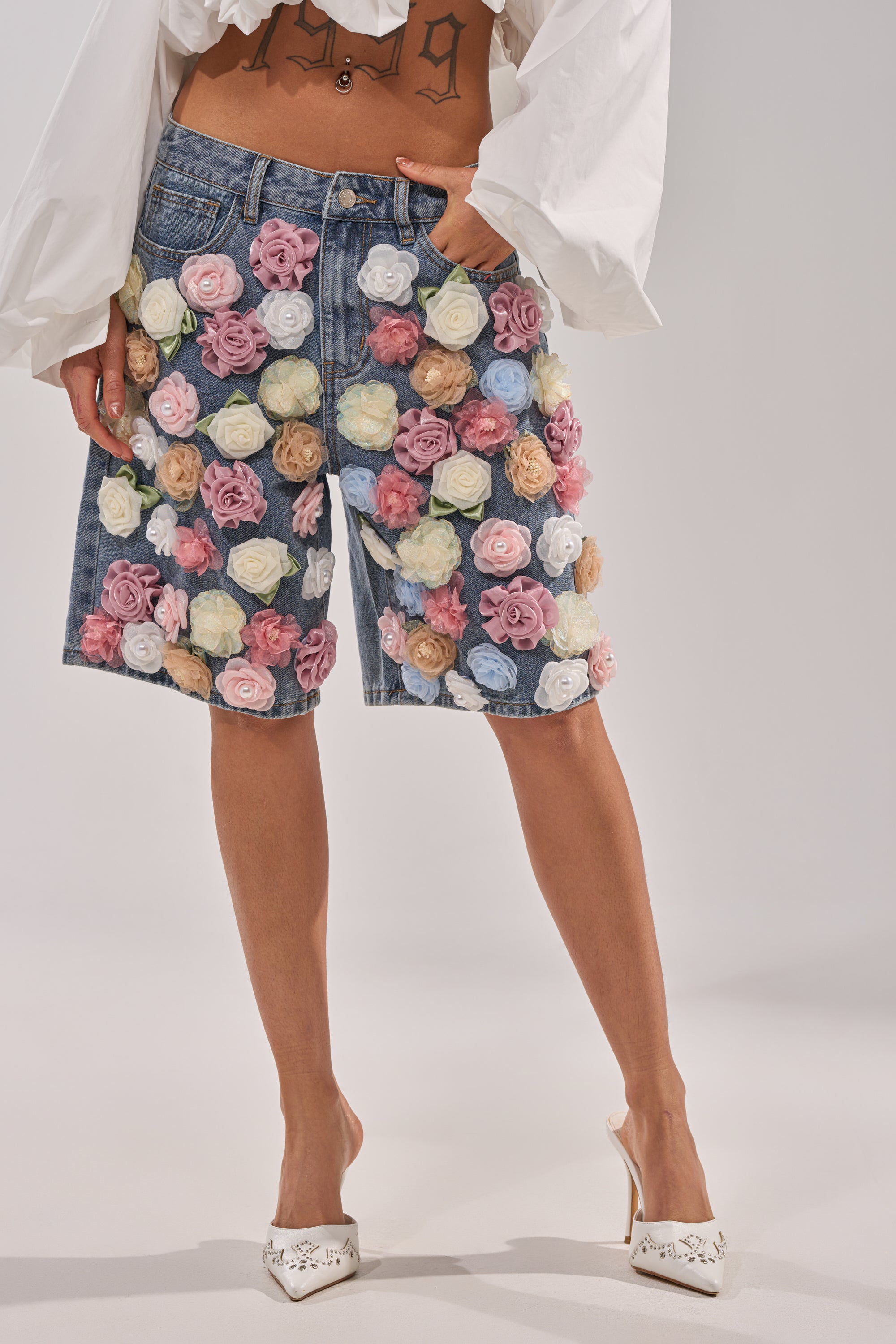 A person wearing the IRIS FLORAL BERMUDA SHORT—high-rise denim shorts with 3D floral appliqués—paired with a white long-sleeve top, white heels, and displaying a number tattoo on their lower abdomen. 2
