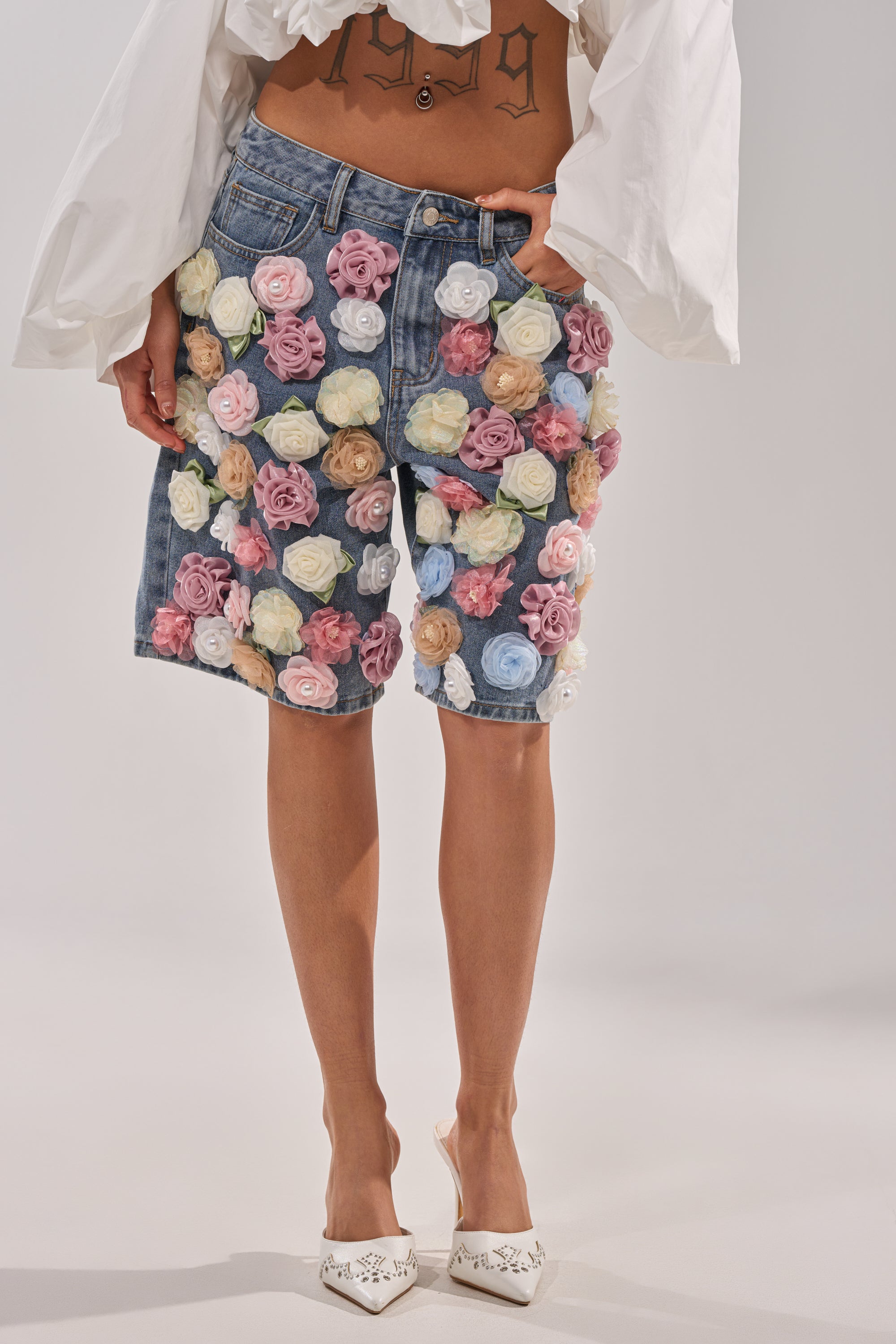 A person is shown from the waist down wearing the IRIS FLORAL BERMUDA SHORT—high-rise denim shorts with 3D floral appliqués—paired with a white blouse and white pointed shoes. 3