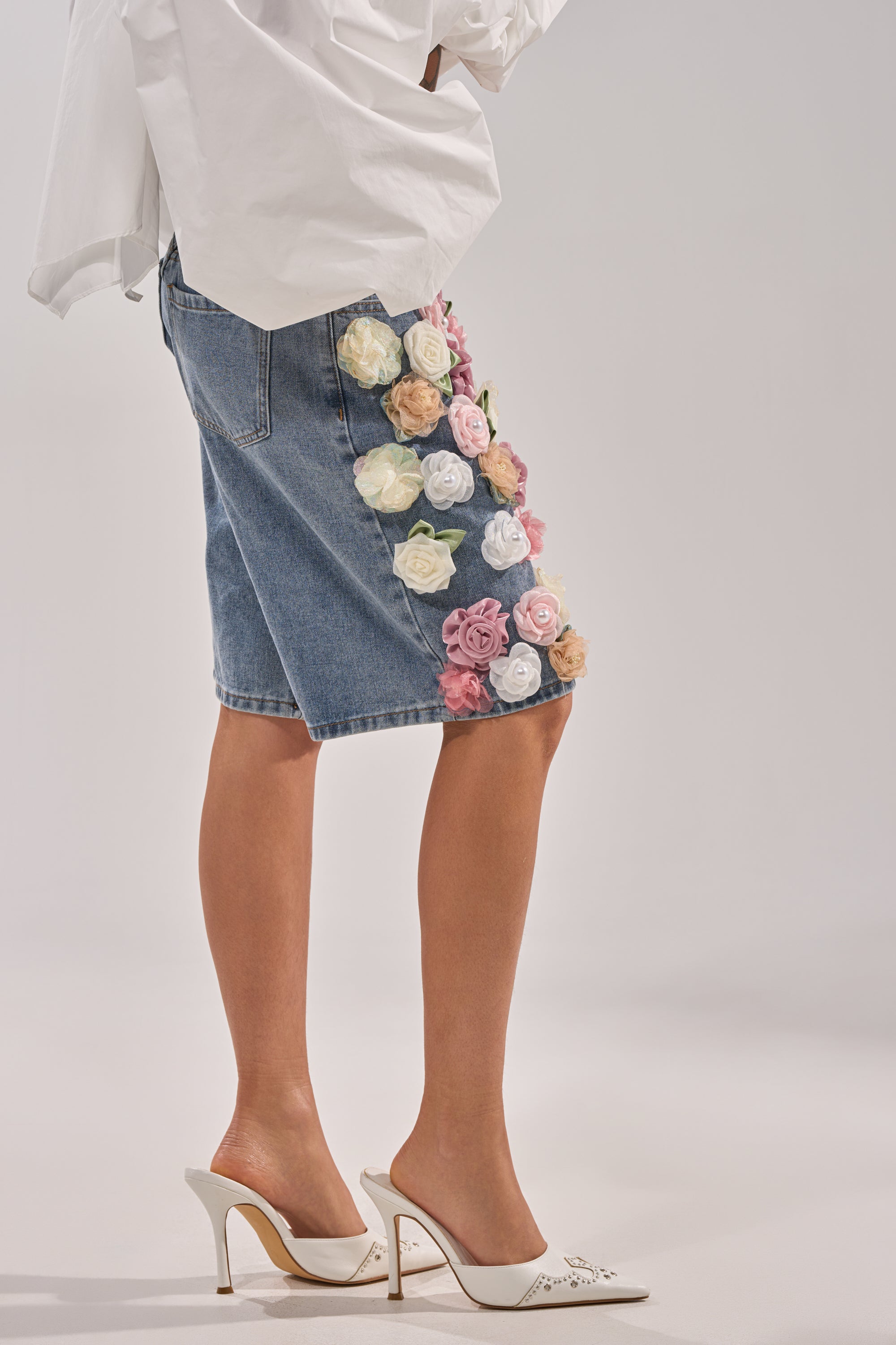 Wearing the IRIS FLORAL BERMUDA SHORT featuring colorful 3D floral appliqués, a person in a white shirt and white heeled mules stands against a plain light background. 4