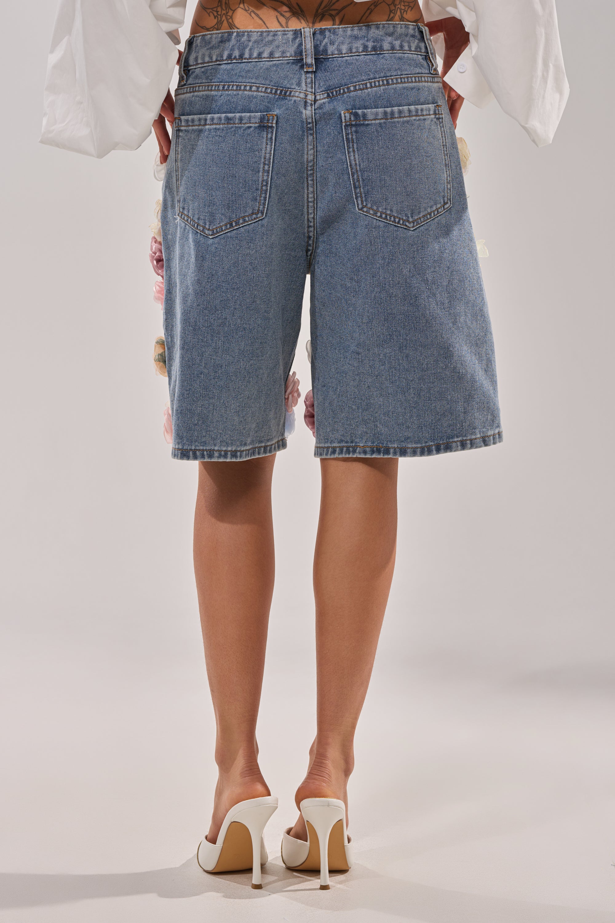 A person stands with their back to the camera against a plain light background, wearing the IRIS FLORAL BERMUDA SHORT and white high-heeled shoes. 5