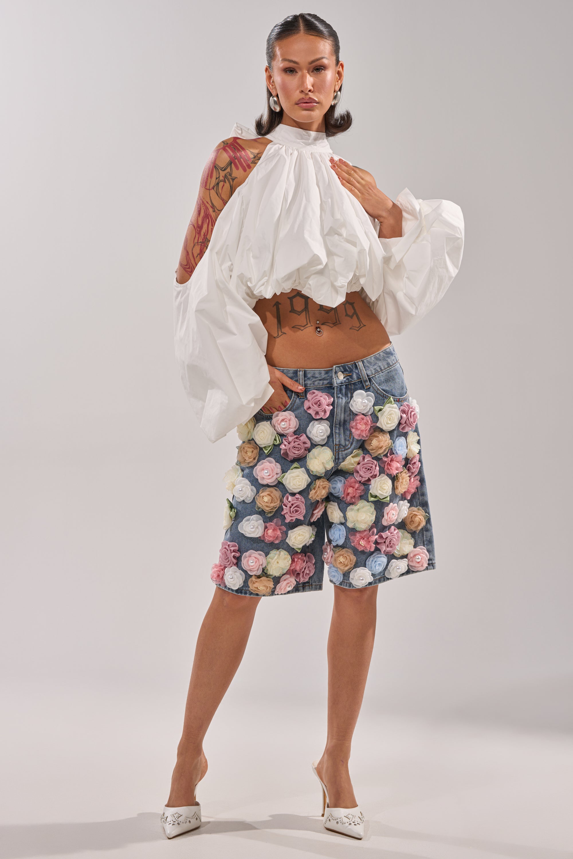 A woman models the IRIS FLORAL BERMUDA SHORT, featuring high-rise denim and 3D floral appliqués, paired with a flowing white blouse with voluminous sleeves. She completes the look with white heels and slicked-back hair. 6