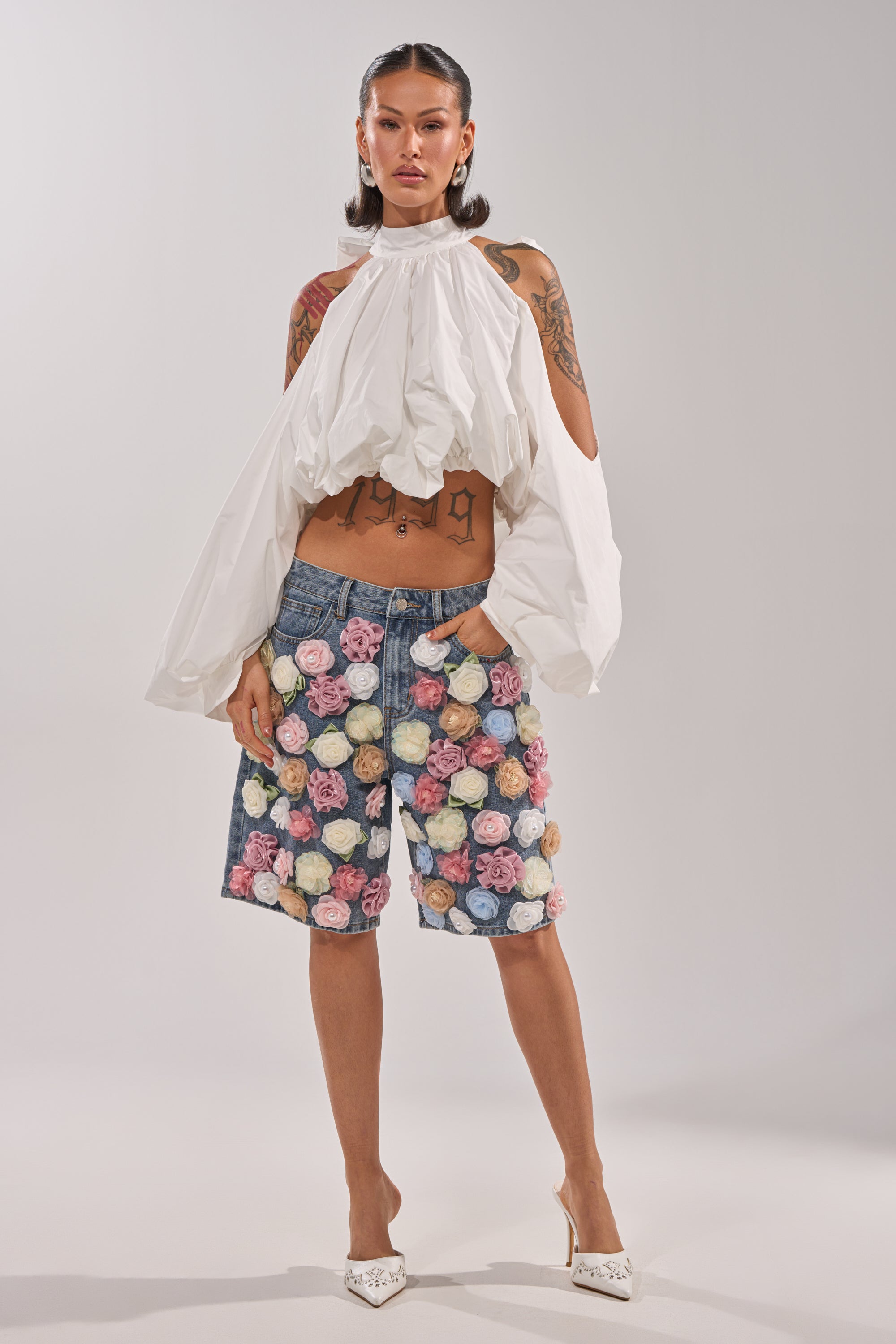 A woman wears the IRIS FLORAL BERMUDA SHORT, a high-rise denim piece with colorful 3D floral appliqués, styled with a white voluminous long-sleeve top, white heels, and showcasing her arm and torso tattoos. 7