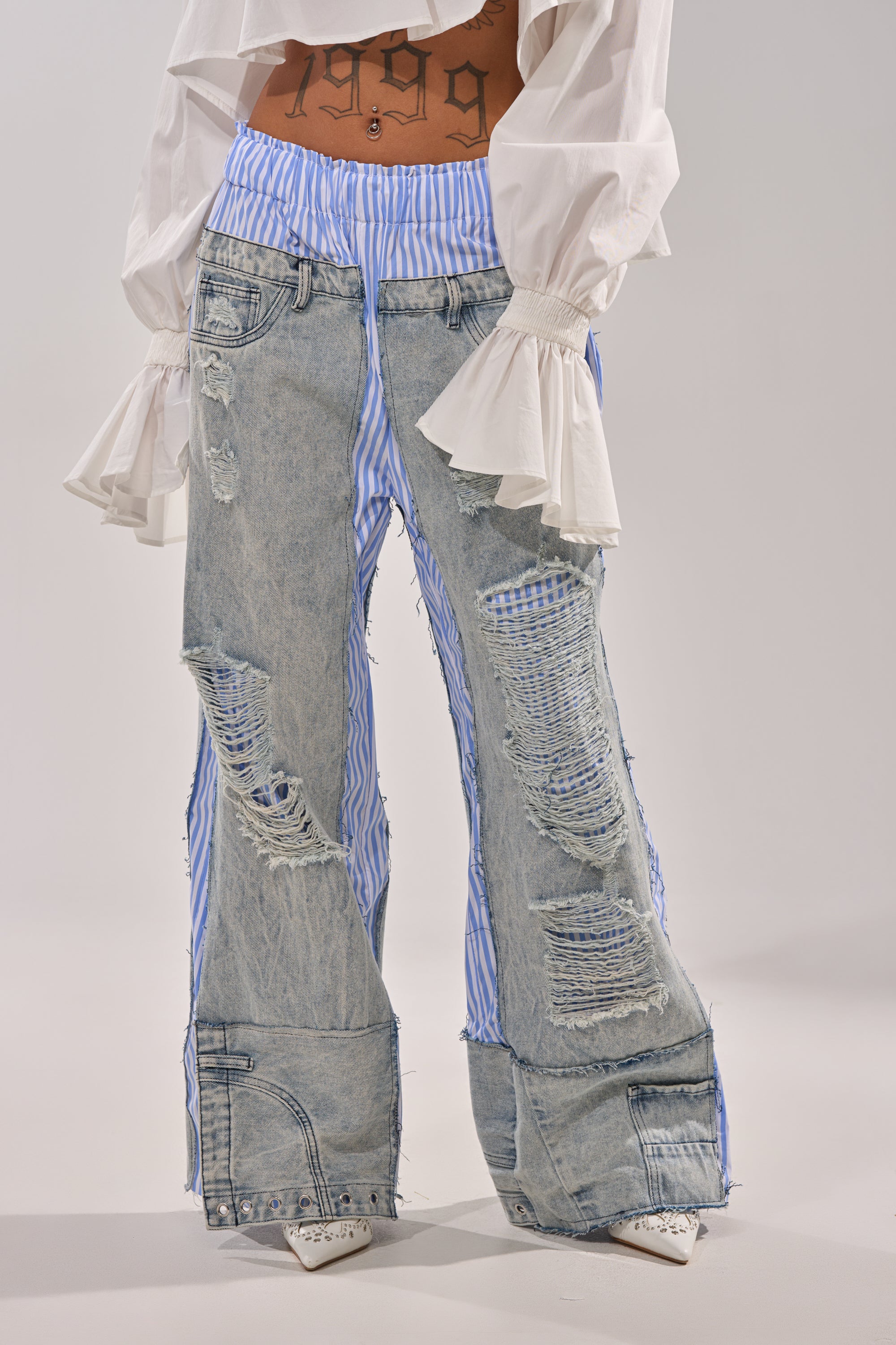 Someone wears the CALL IT WHAT YOU WANT MIXED UP TROUSER—distressed, light-wash denim with blue-and-white striped inserts and frayed hems—with a white ruffled blouse and pointed shoes, revealing a "1999" tattoo at the midriff. 0