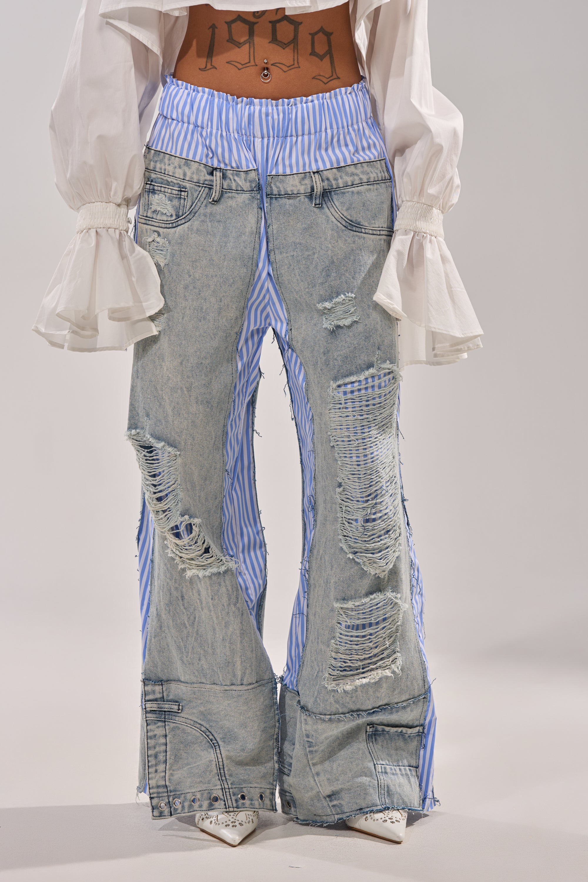 Wearing the CALL IT WHAT YOU WANT MIXED UP TROUSER, featuring blue-and-white boxer stripe details, styled with a white flared-sleeve blouse and a "1999" tattoo at the midriff for a striking street-style look. 1