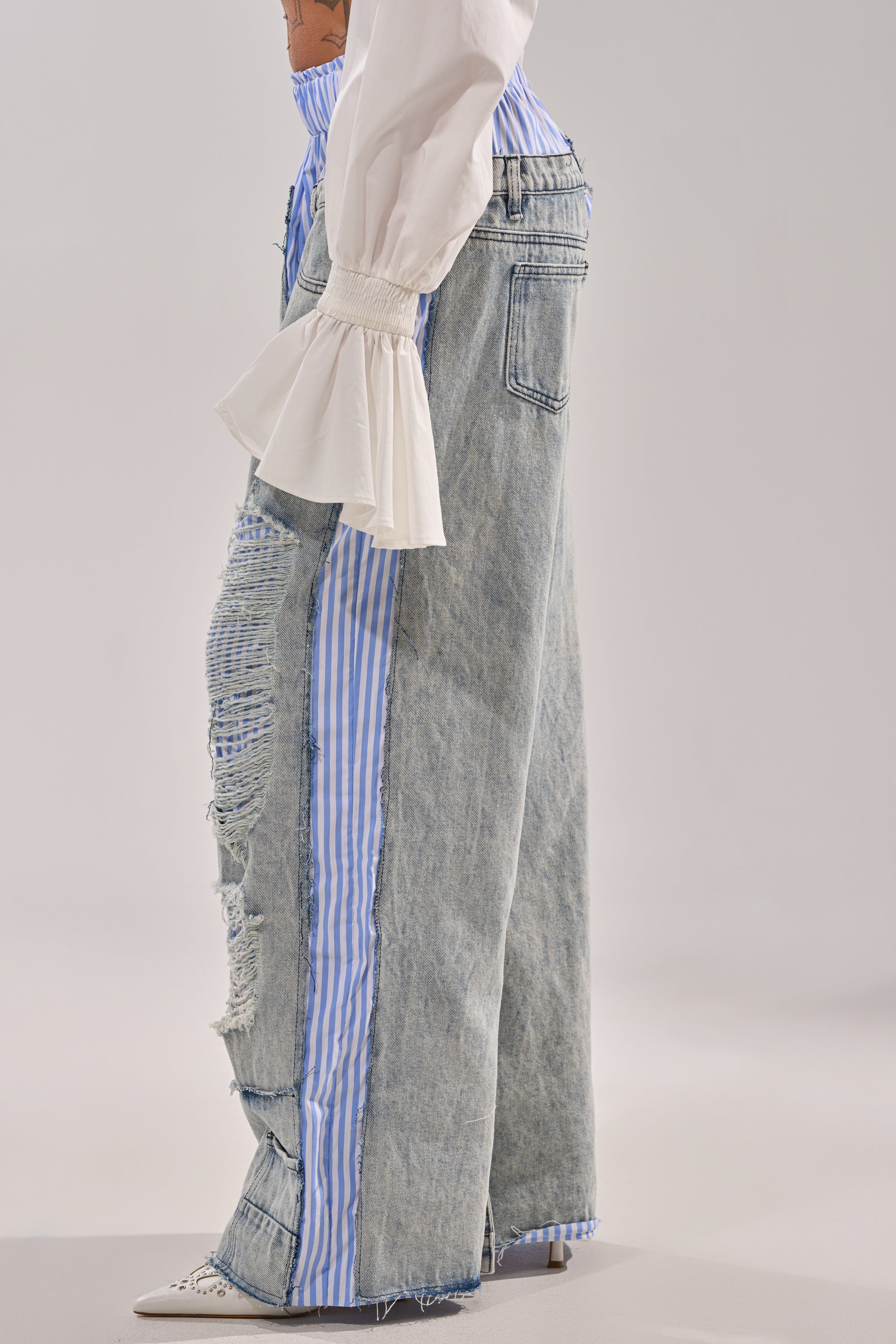 Wearing the CALL IT WHAT YOU WANT MIXED UP TROUSER—wide-leg, distressed denim with blue and white striped inserts—styled with a white blouse with flared ruffled cuffs and pointed-toe white heels. 3