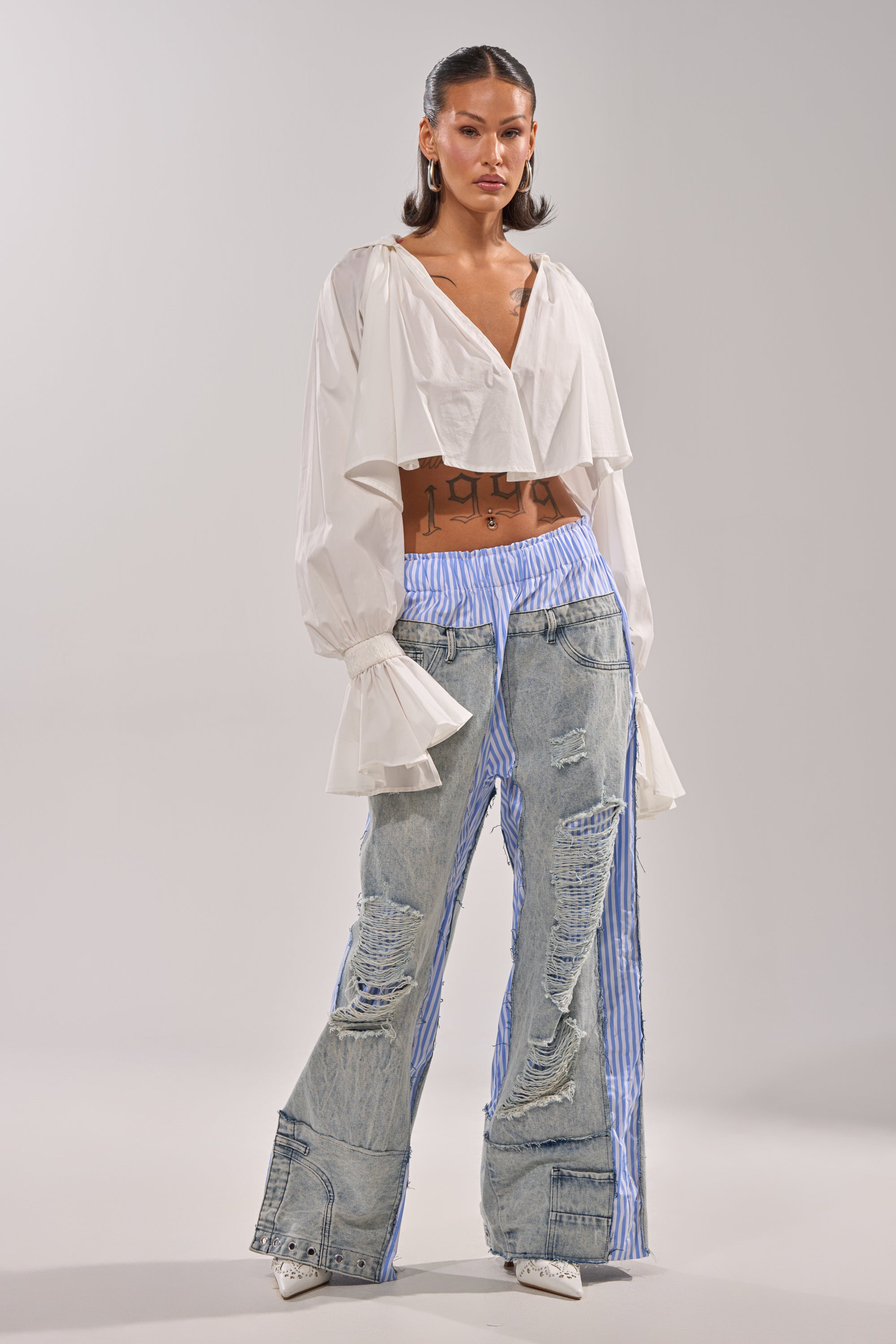 A woman wears a white ruffled crop top and the CALL IT WHAT YOU WANT MIXED UP TROUSER—high-waisted, wide-leg jeans with distressed patches and blue-and-white pinstripes—paired with hoop earrings and pointed white shoes. 5