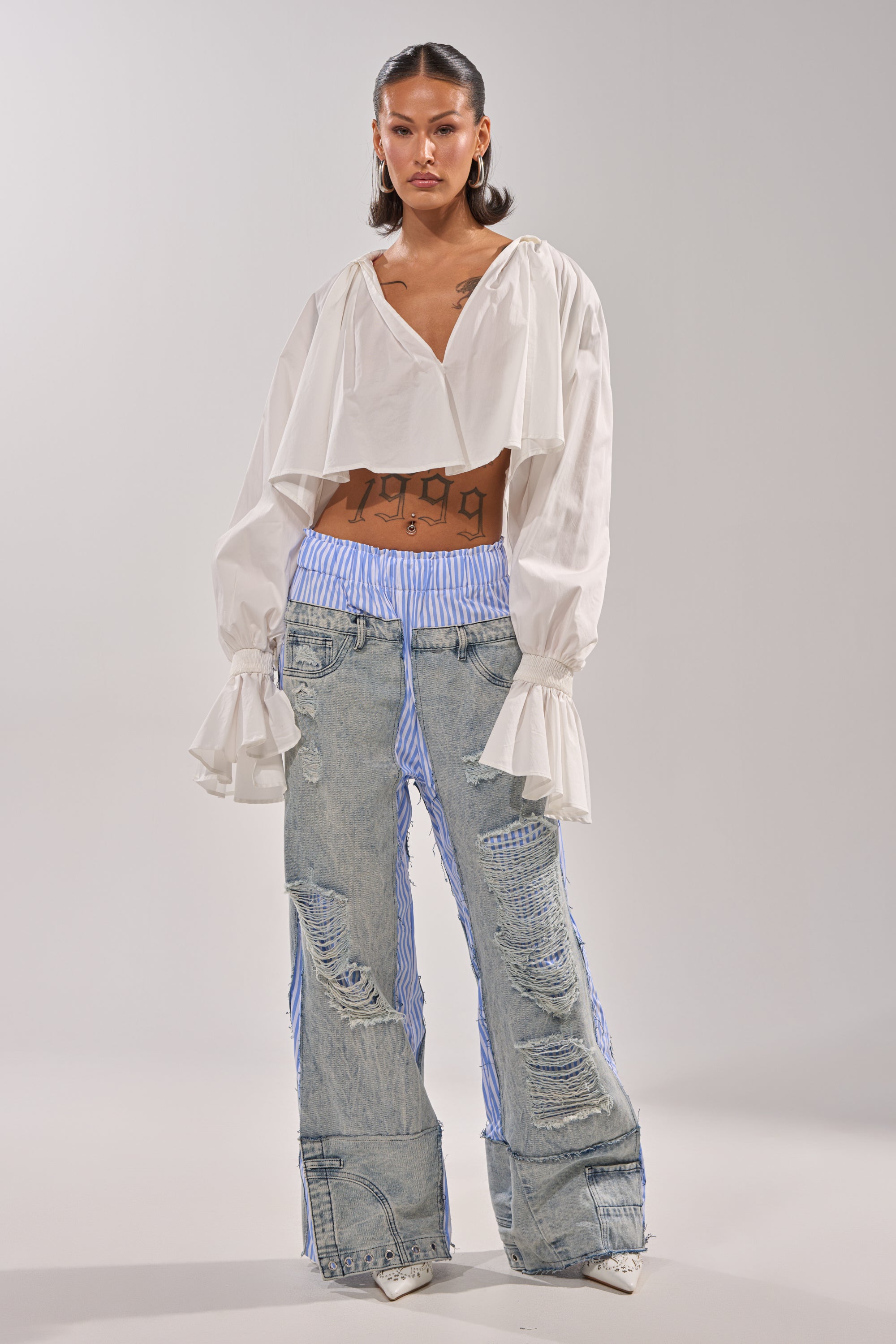 A woman stands against a plain background, wearing a white, wide-sleeved, cropped blouse and the CALL IT WHAT YOU WANT MIXED UP TROUSER—hybrid denim pants featuring blue pinstripe fabric beneath. Her hair is styled back and she has torso tattoos. 6