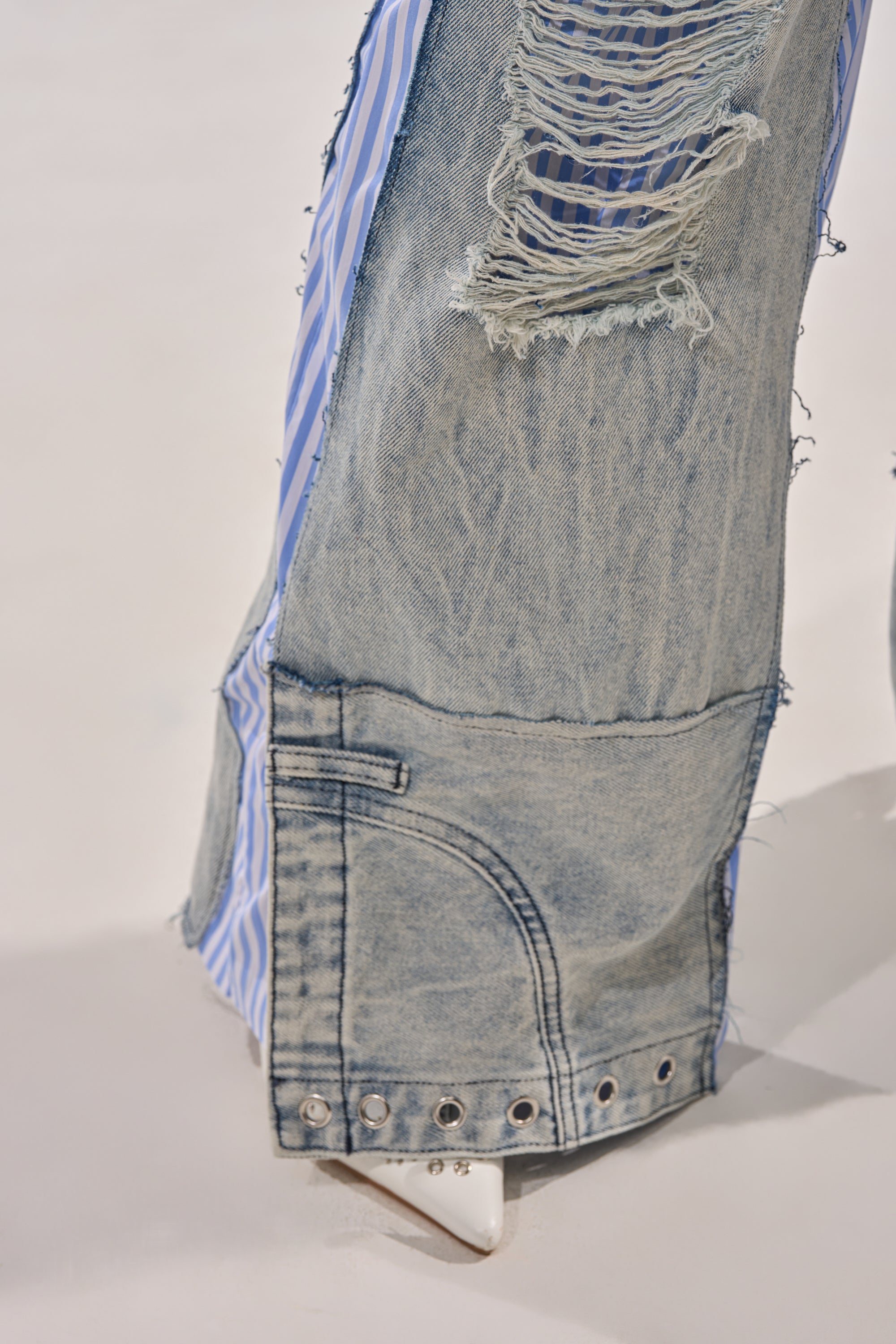 Close-up of the CALL IT WHAT YOU WANT MIXED UP TROUSER—street-style hybrid denim pants with frayed edges, blue and white boxer stripe details underneath, silver hem rivets, and a peek of a white pointed-toe shoe. 7