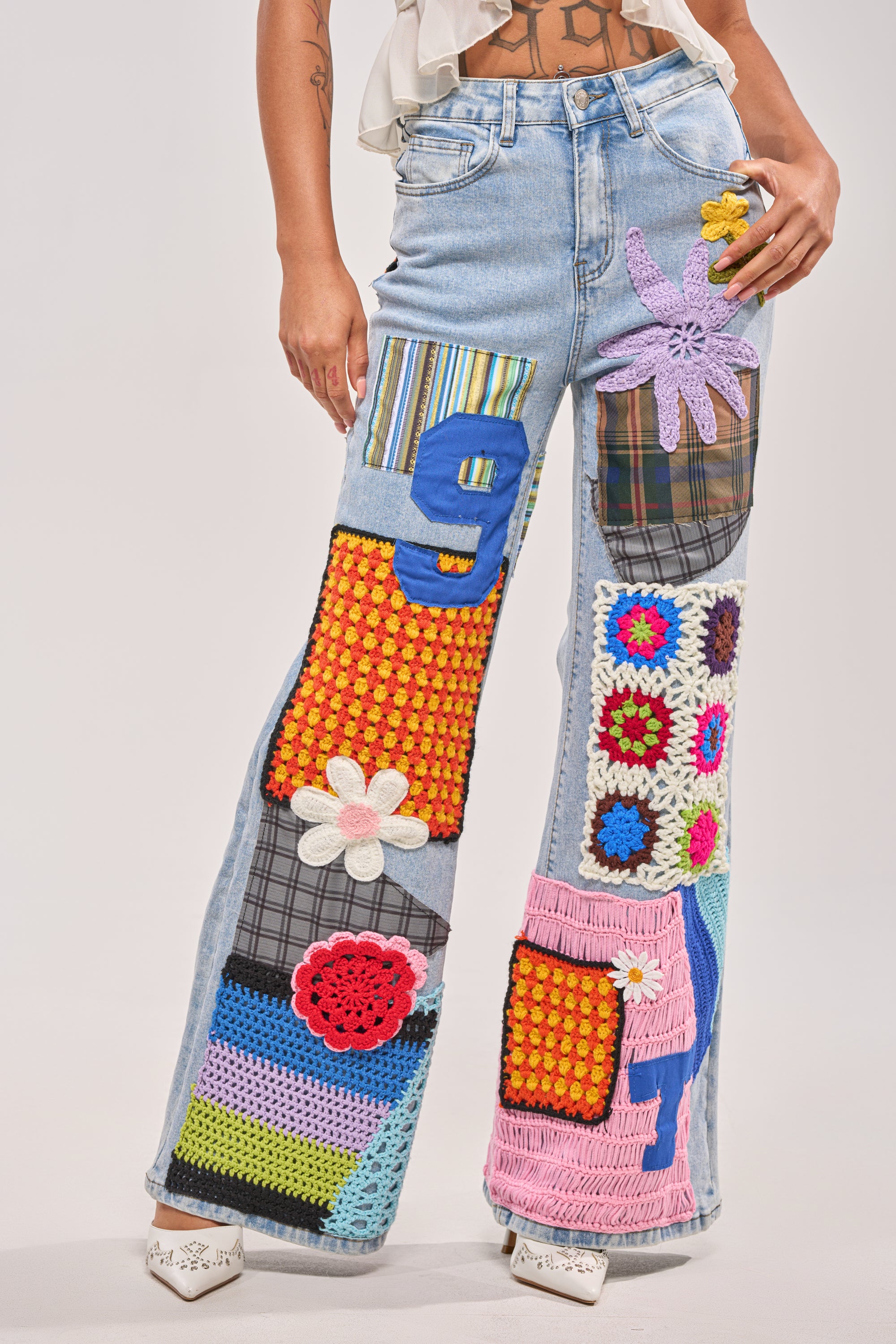 Wearing CUTE LIKE ME RELAXED DENIM wide-leg jeans in light blue, featuring crochet patchwork with flowers, numbers, and textured fabrics, paired with white heeled shoes for a playful street style vibe. Only the lower half is visible. 0