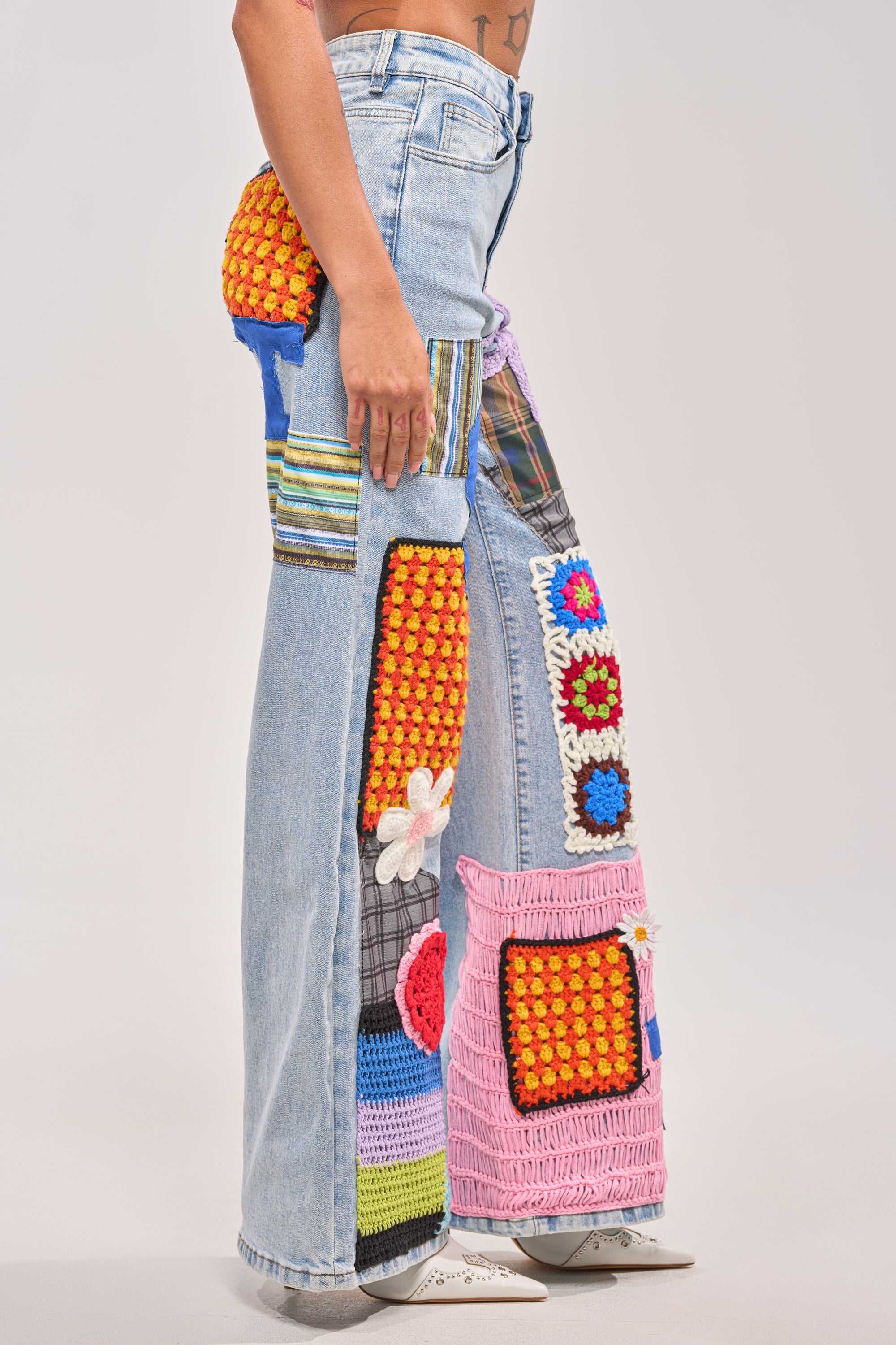 A person wearing the CUTE LIKE ME RELAXED DENIM, featuring light blue crochet patchwork and colorful fabric patches in different patterns, stands against a plain backdrop—only their lower torso and legs are visible, highlighting unique street style. 2