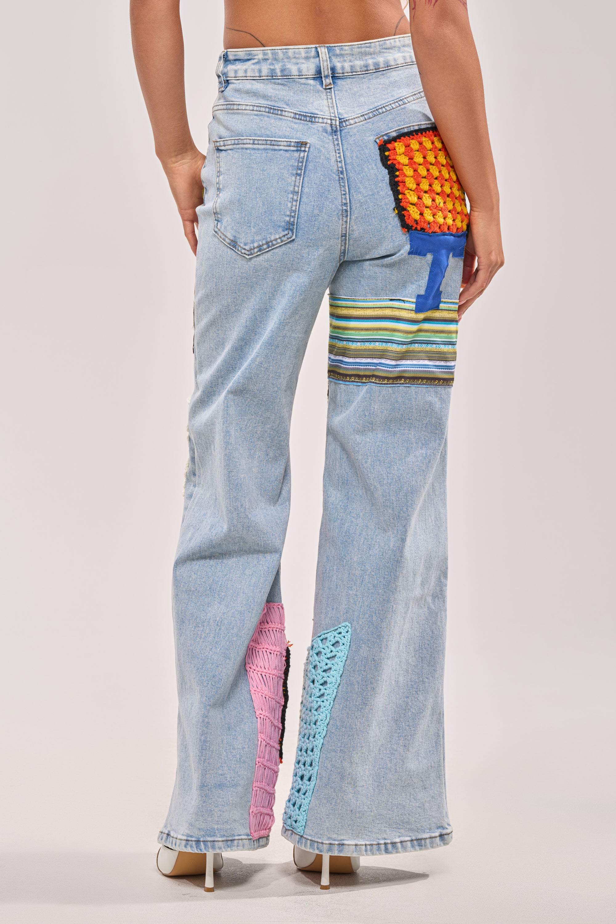Captured from behind, someone wears the CUTE LIKE ME RELAXED DENIM—light blue, high-waisted crochet patchwork jeans with colorful patches and side/back stripes—styled with white heels for an effortless street vibe. 3