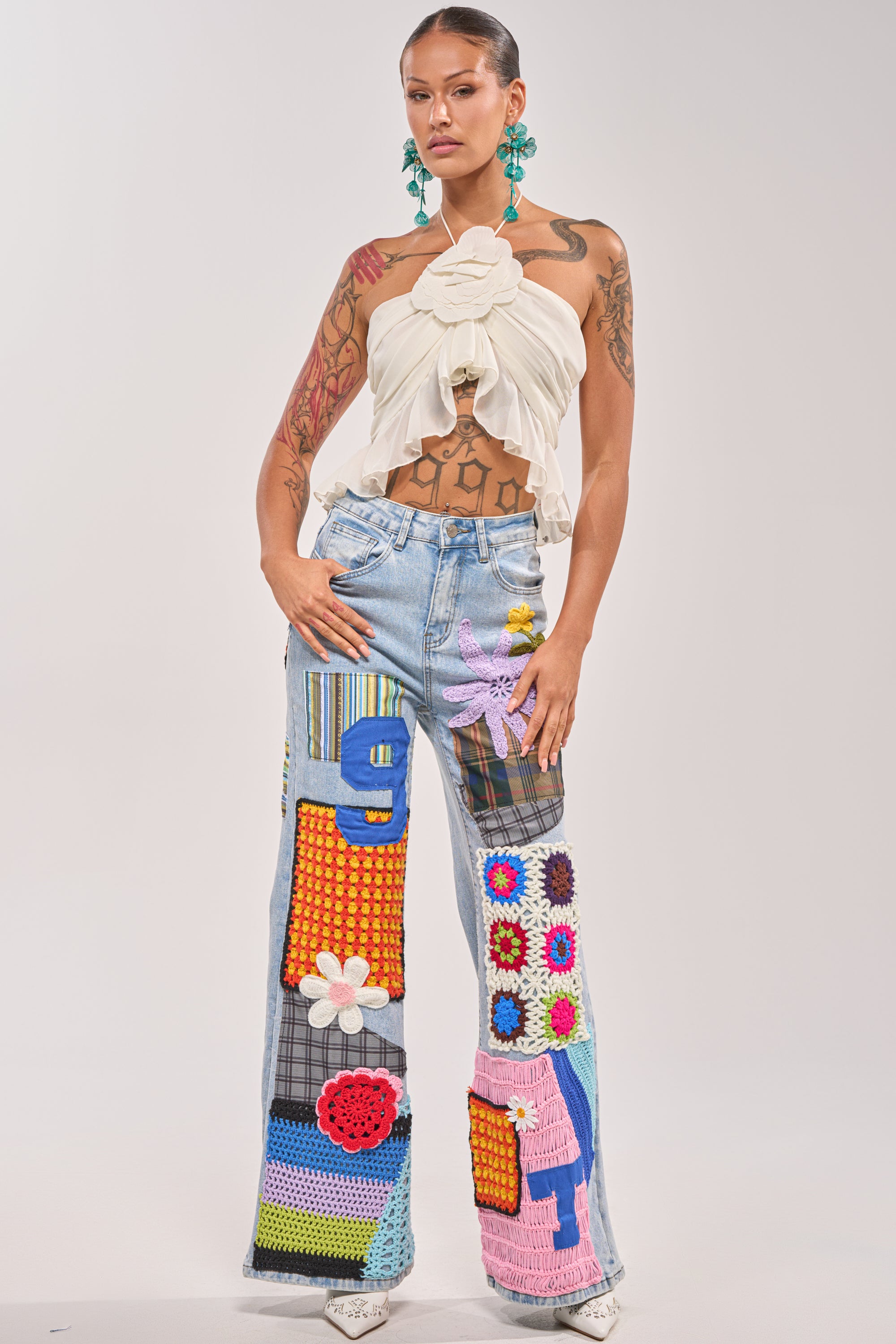 A person exudes vibrant street style in the CUTE LIKE ME RELAXED DENIM—colorful crochet patchwork wide-leg jeans with bold flower designs—paired with a white floral halter top, turquoise earrings, and visible arm and chest tattoos. 4