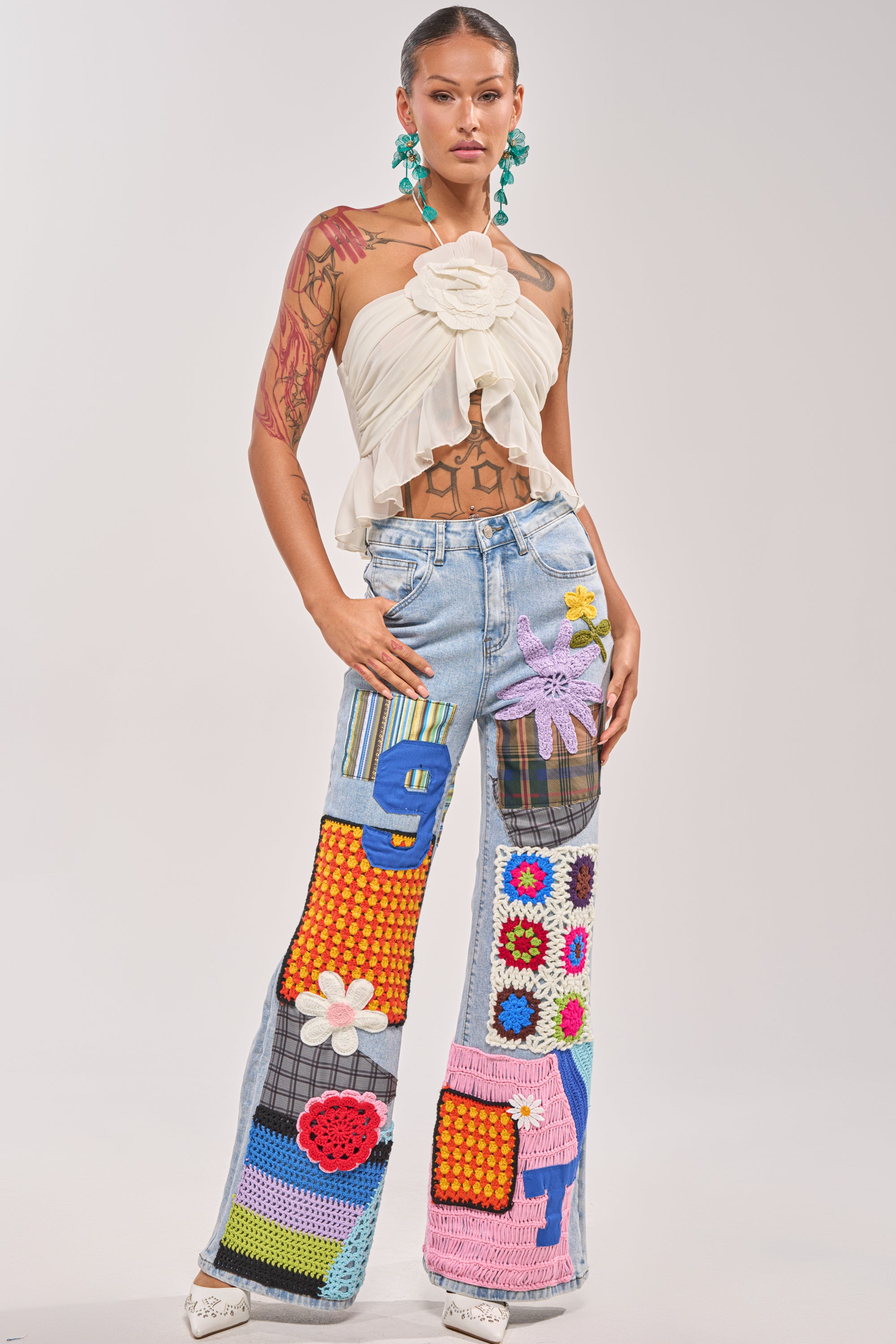 A person poses confidently in the CUTE LIKE ME RELAXED DENIM, street style patchwork jeans with numbers, flowers, and patterns, paired with a white halter top featuring a large flower detail. They accessorize with teal earrings and display their tattoos. 5