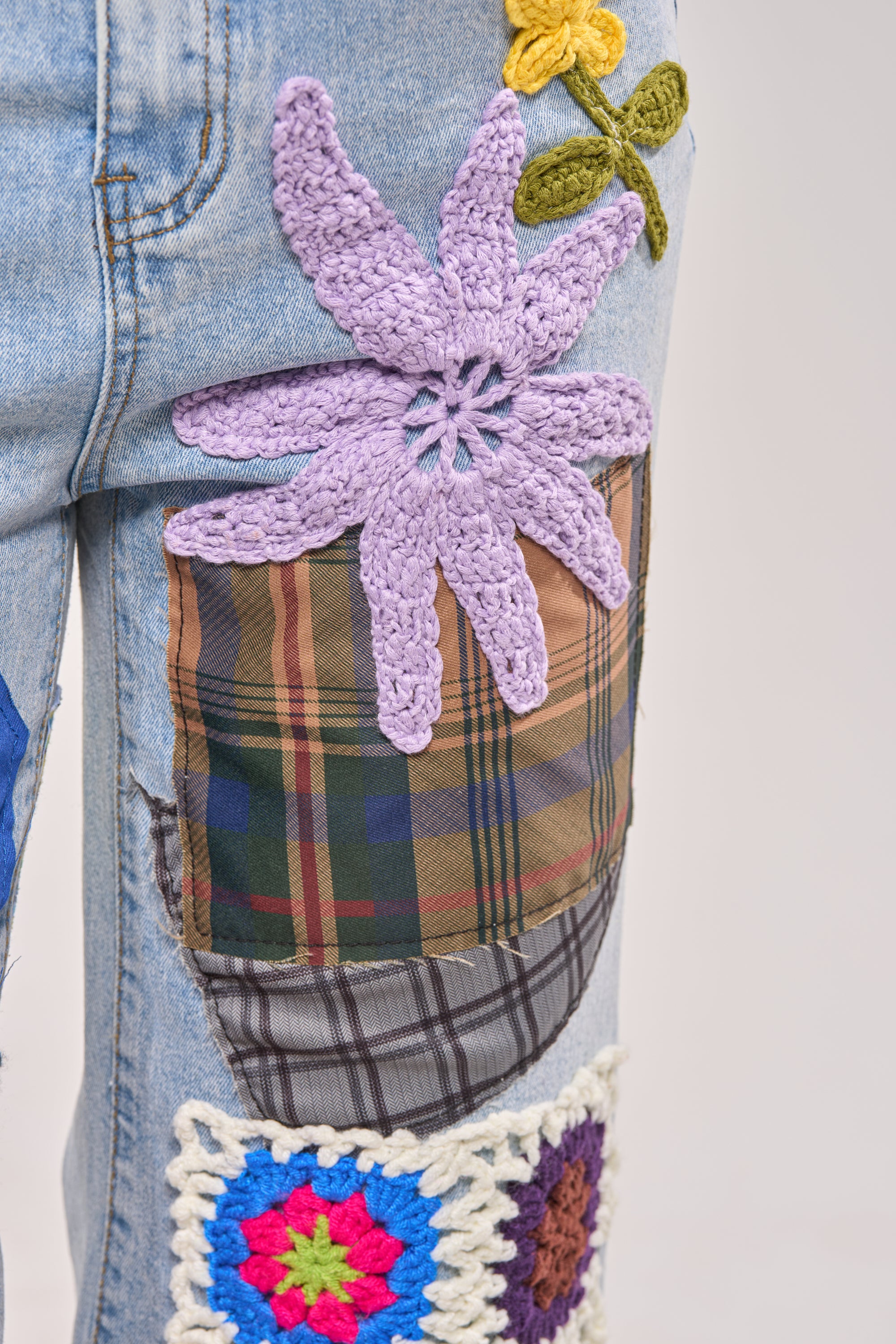 Close-up of CUTE LIKE ME RELAXED DENIM jeans with crochet patchwork, including colorful flower patches, a large lilac crocheted flower, a plaid rectangle, and multicolored squares for unique street style. 6