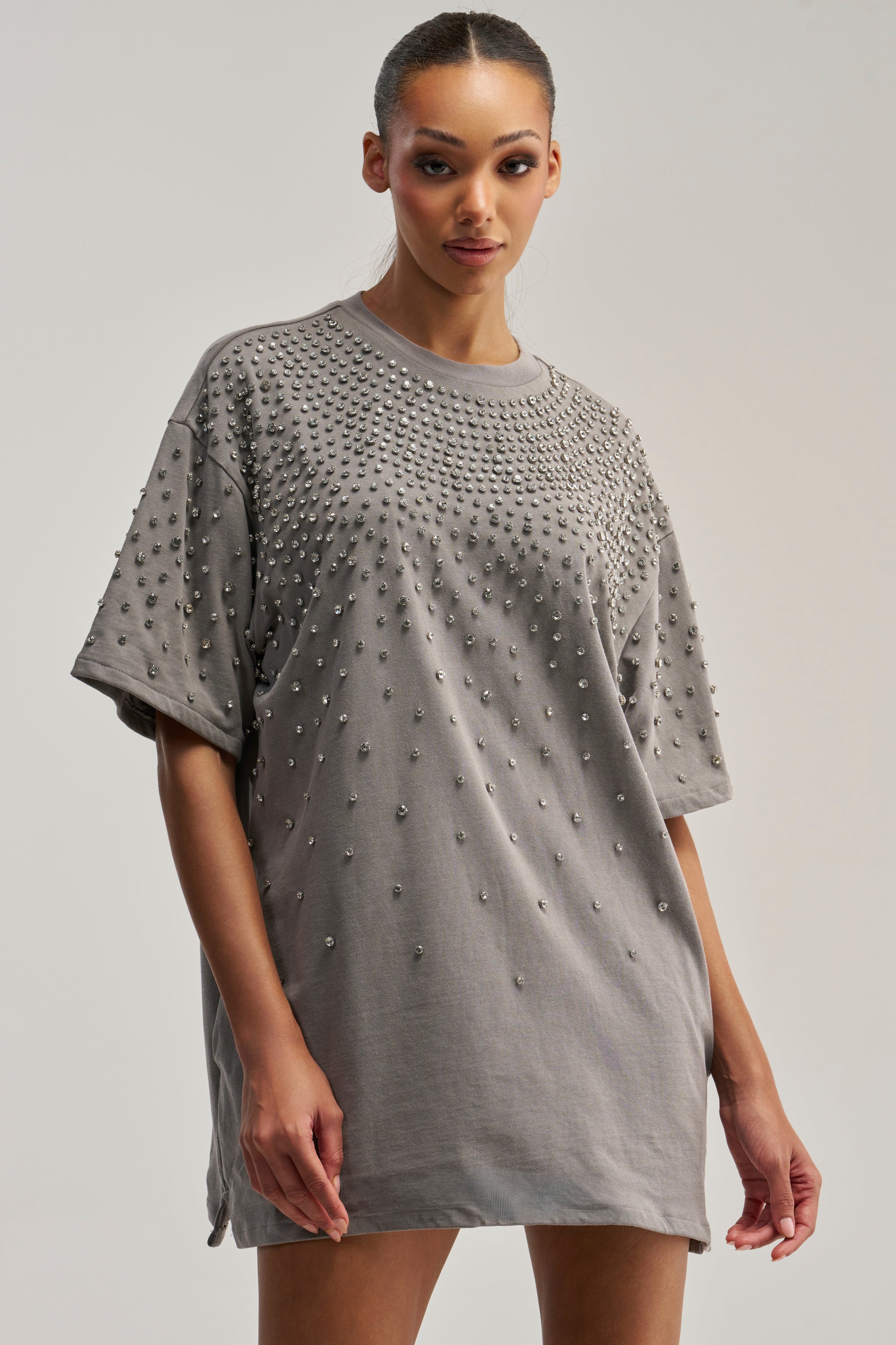 A woman models the IN MY ELEMENT RHINESTONE TEE SHIRT MINI DRESS, a loose gray dress embellished with small metal studs across the shoulders and chest. She stands against a plain background, gazing confidently at the camera.