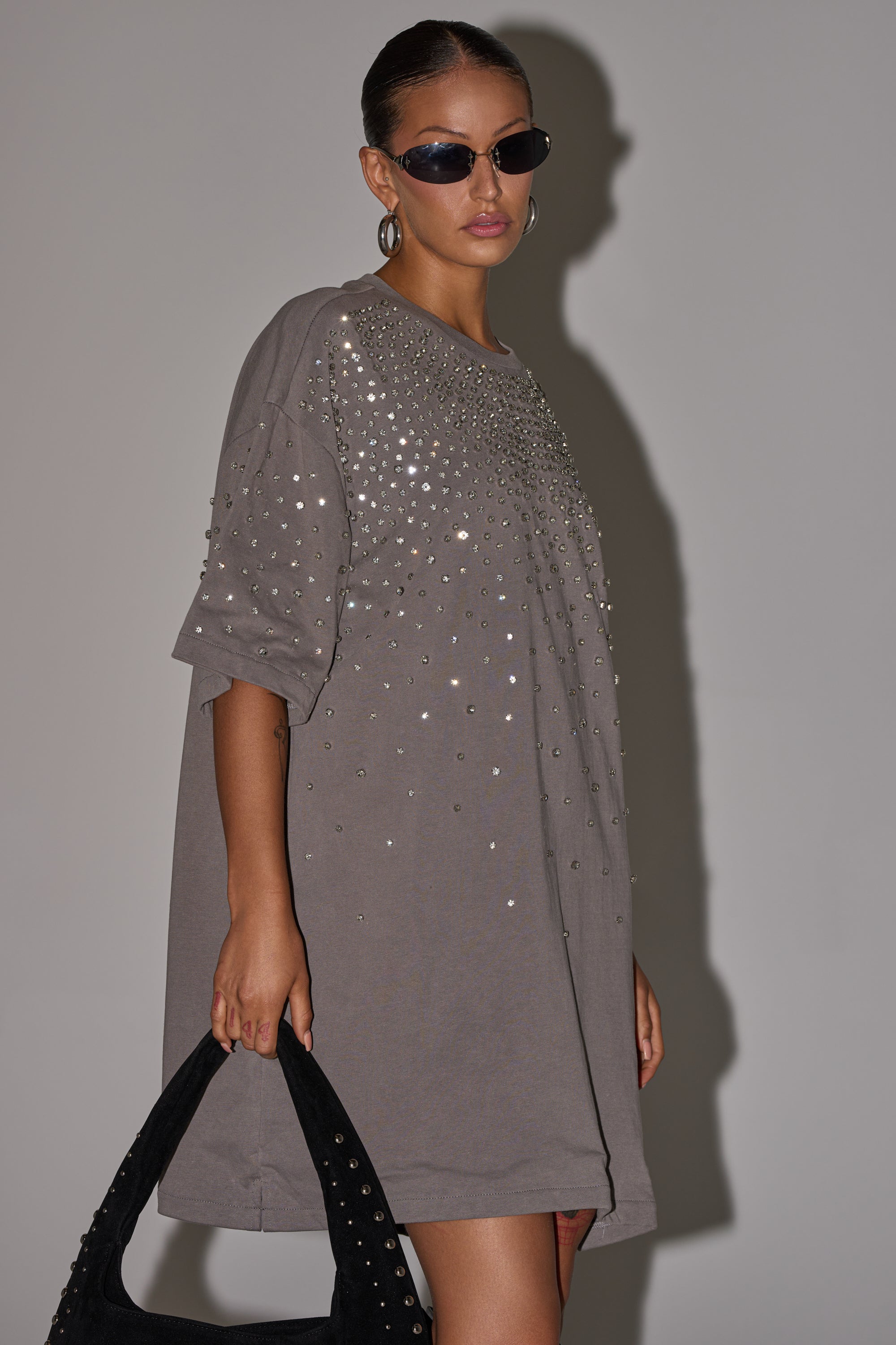 A woman in the IN MY ELEMENT RHINESTONE TEE SHIRT MINI DRESS, styled with sunglasses and hoop earrings, holds a black studded handbag against a neutral background. 1