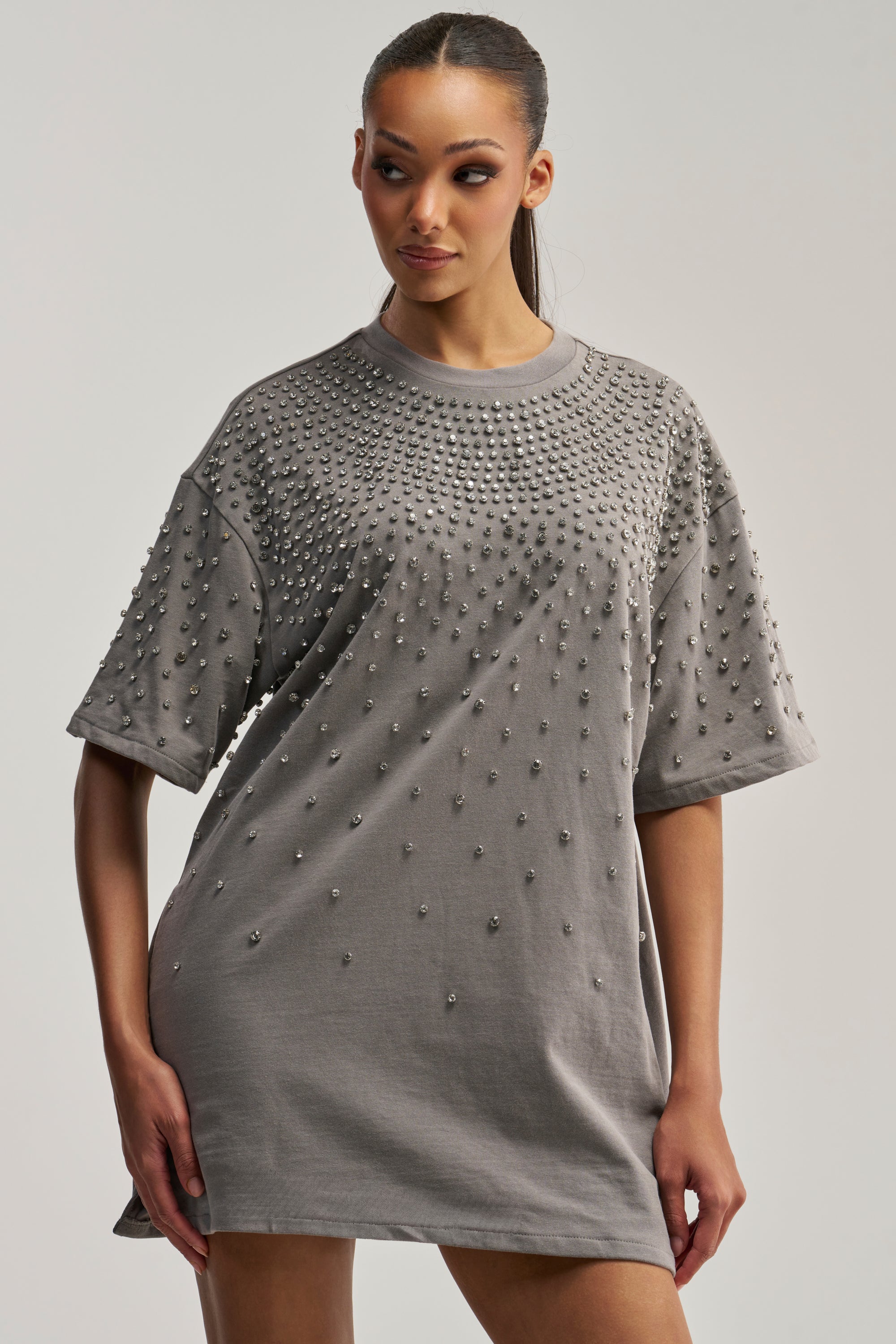 A woman stands against a plain light background, looking to her left, wearing the IN MY ELEMENT RHINESTONE TEE SHIRT MINI DRESS—an oversized grey T-shirt dress embellished with numerous small, round crystal rhinestones on the front and sleeves.