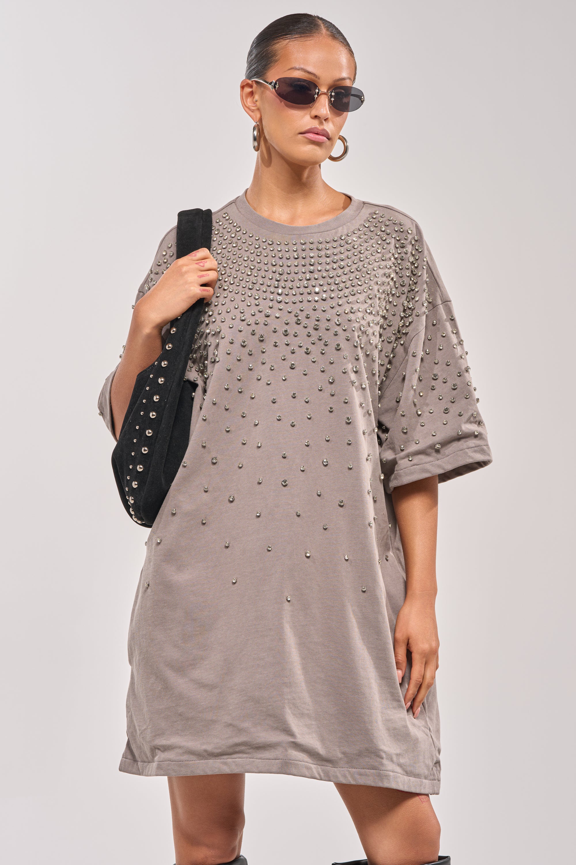 A woman stands confidently against a neutral backdrop in the IN MY ELEMENT RHINESTONE TEE SHIRT MINI DRESS, paired with sunglasses, hoop earrings, and a black studded shoulder bag. 2