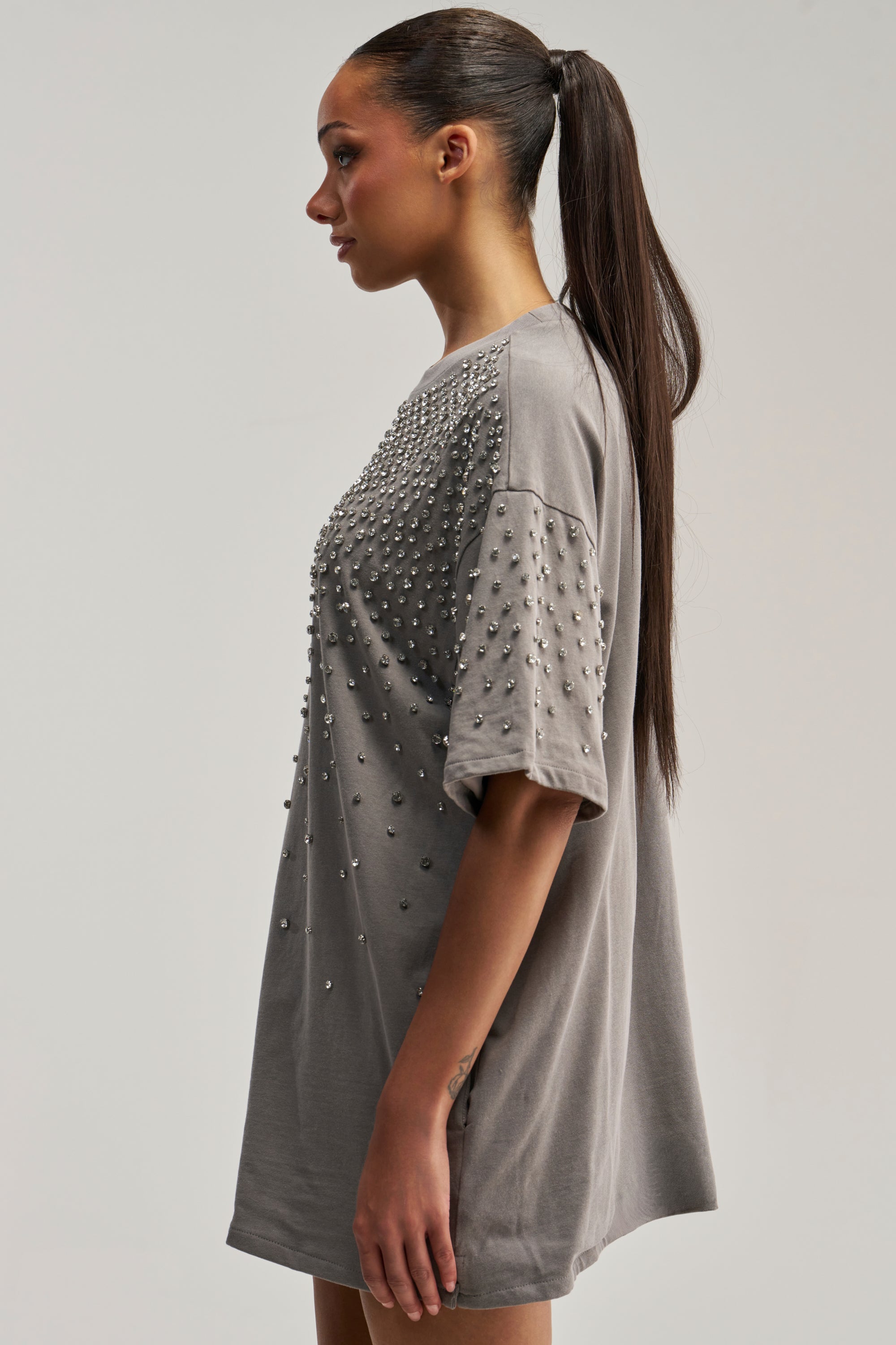 A woman in profile with a long ponytail showcases the IN MY ELEMENT RHINESTONE TEE SHIRT MINI DRESS, an oversized grey dress embellished with small rhinestones on the front and sleeves, standing against a plain background.