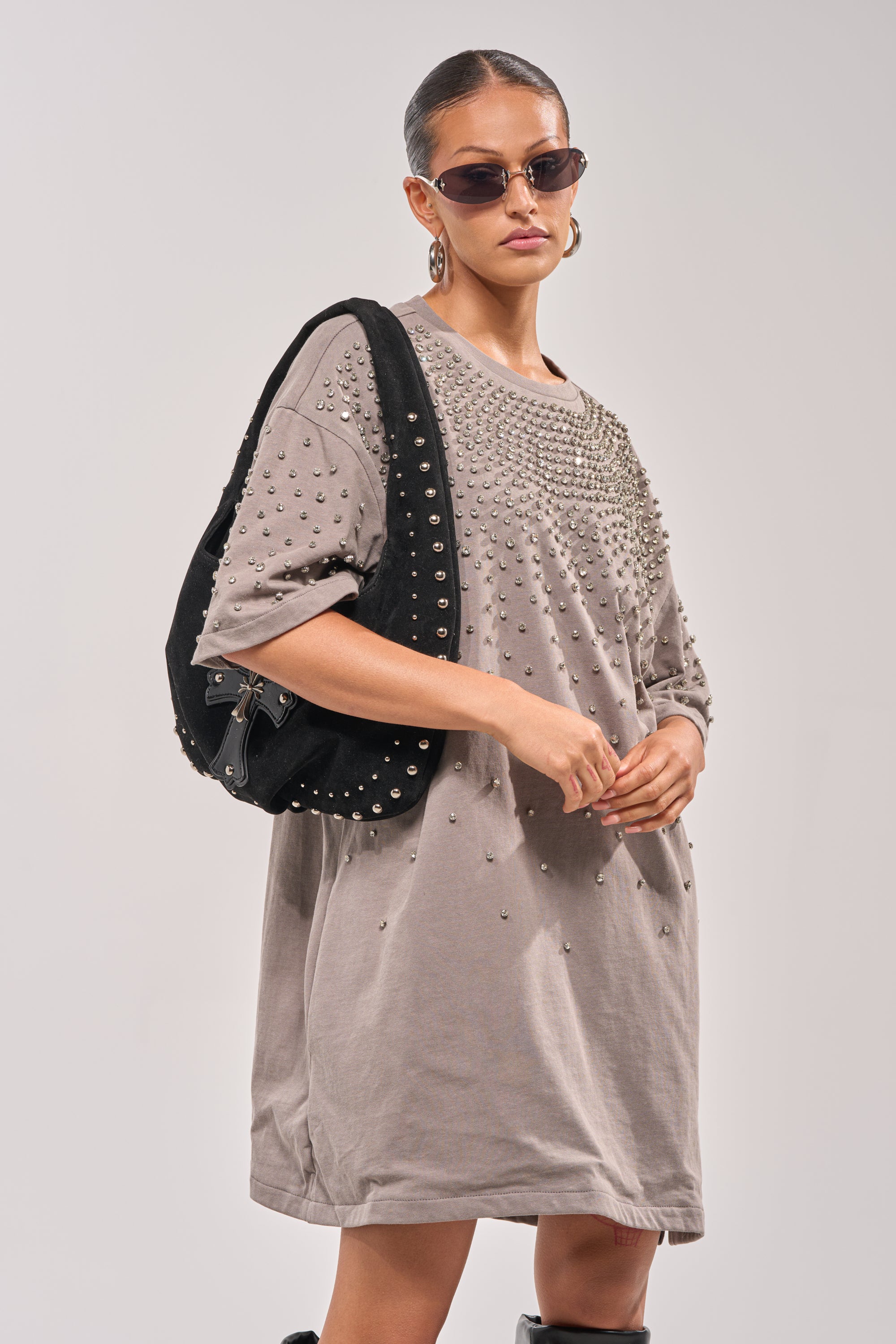 A woman stands confidently in the IN MY ELEMENT RHINESTONE TEE SHIRT MINI DRESS, accessorized with sunglasses, hoop earrings, a black studded bag, her hair pulled back, and knee-high black boots for a chic look. 3