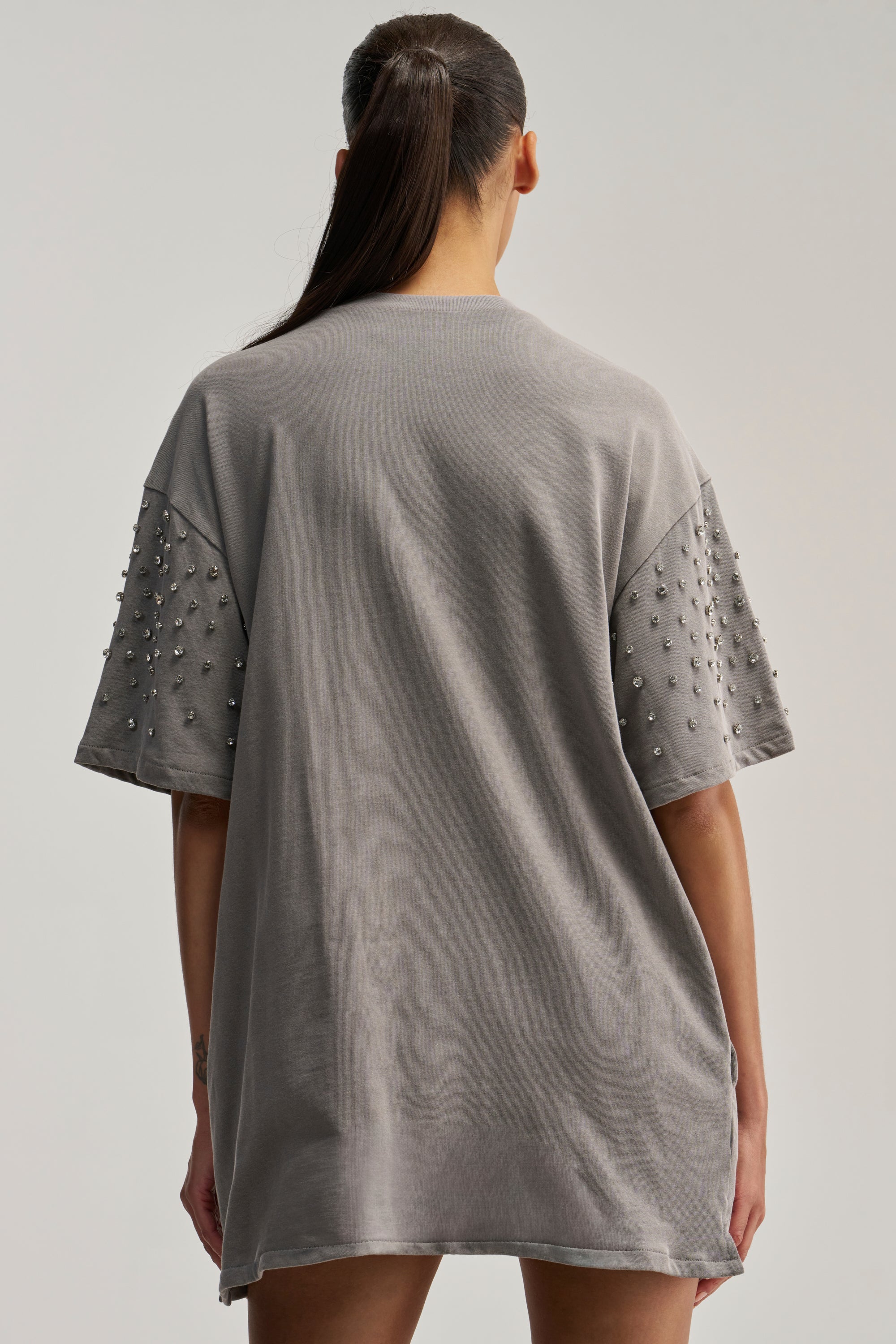 A person with long dark hair in a ponytail is shown from the back against a plain light background, wearing the IN MY ELEMENT RHINESTONE TEE SHIRT MINI DRESS—an oversized grey dress featuring decorative rhinestone studs on the sleeves.