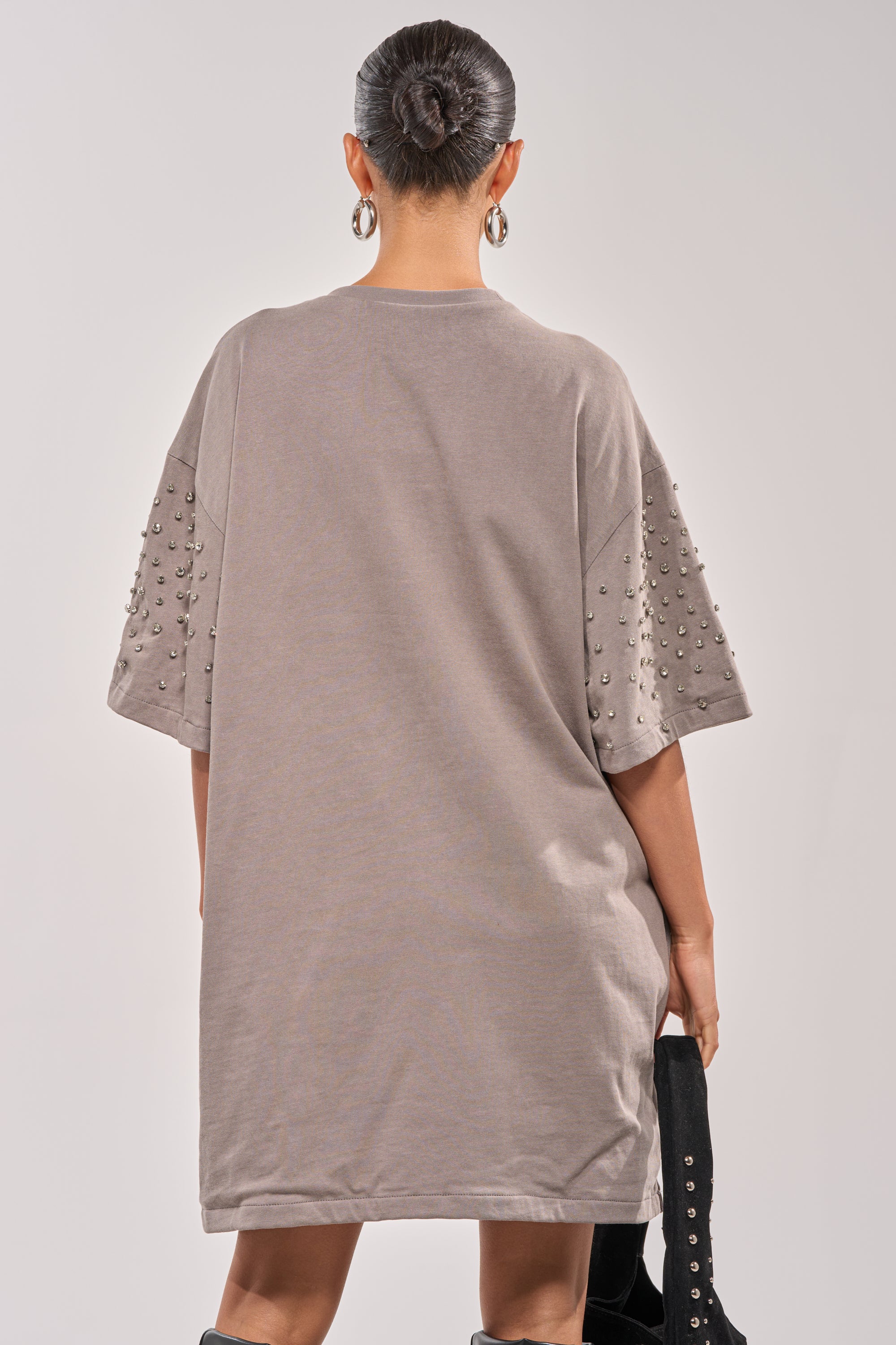 A person with a sleek bun is seen from behind wearing the IN MY ELEMENT RHINESTONE TEE SHIRT MINI DRESS, featuring metallic studs on the sleeves, paired with knee-high black boots, and holding a black garment. 5