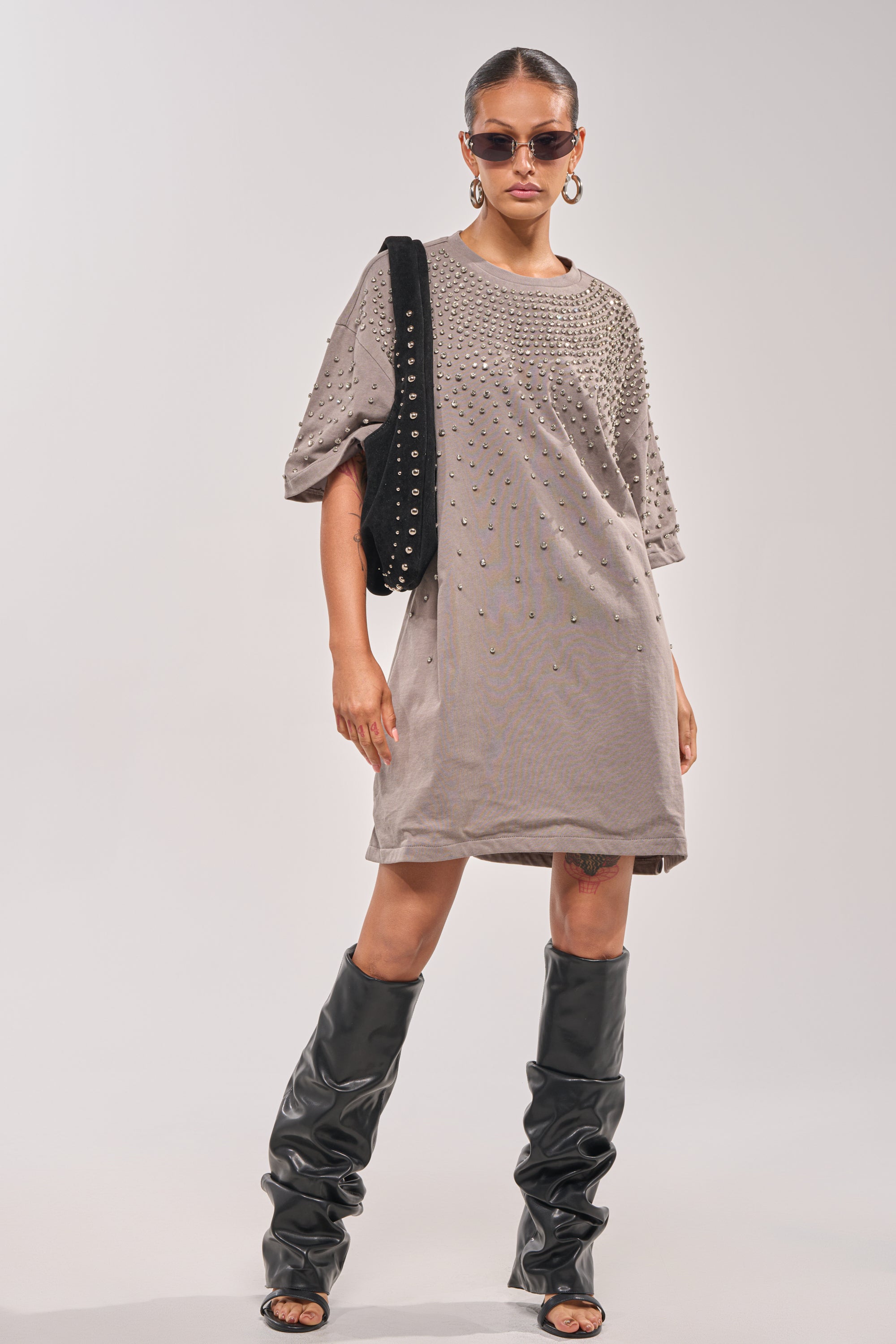 A woman stands confidently against a plain background wearing the IN MY ELEMENT RHINESTONE TEE SHIRT MINI DRESS, paired with black knee-high boots, hoop earrings, sunglasses, and carrying a black studded shoulder bag. 6