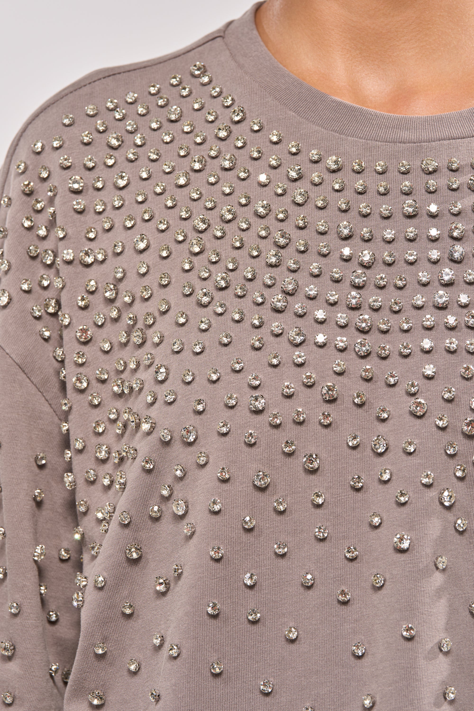 Close-up of the IN MY ELEMENT RHINESTONE TEE SHIRT MINI DRESS in taupe, featuring a crewneck and rows of shiny silver studs across the fabric. The neckline and shoulder seam are visible. 7
